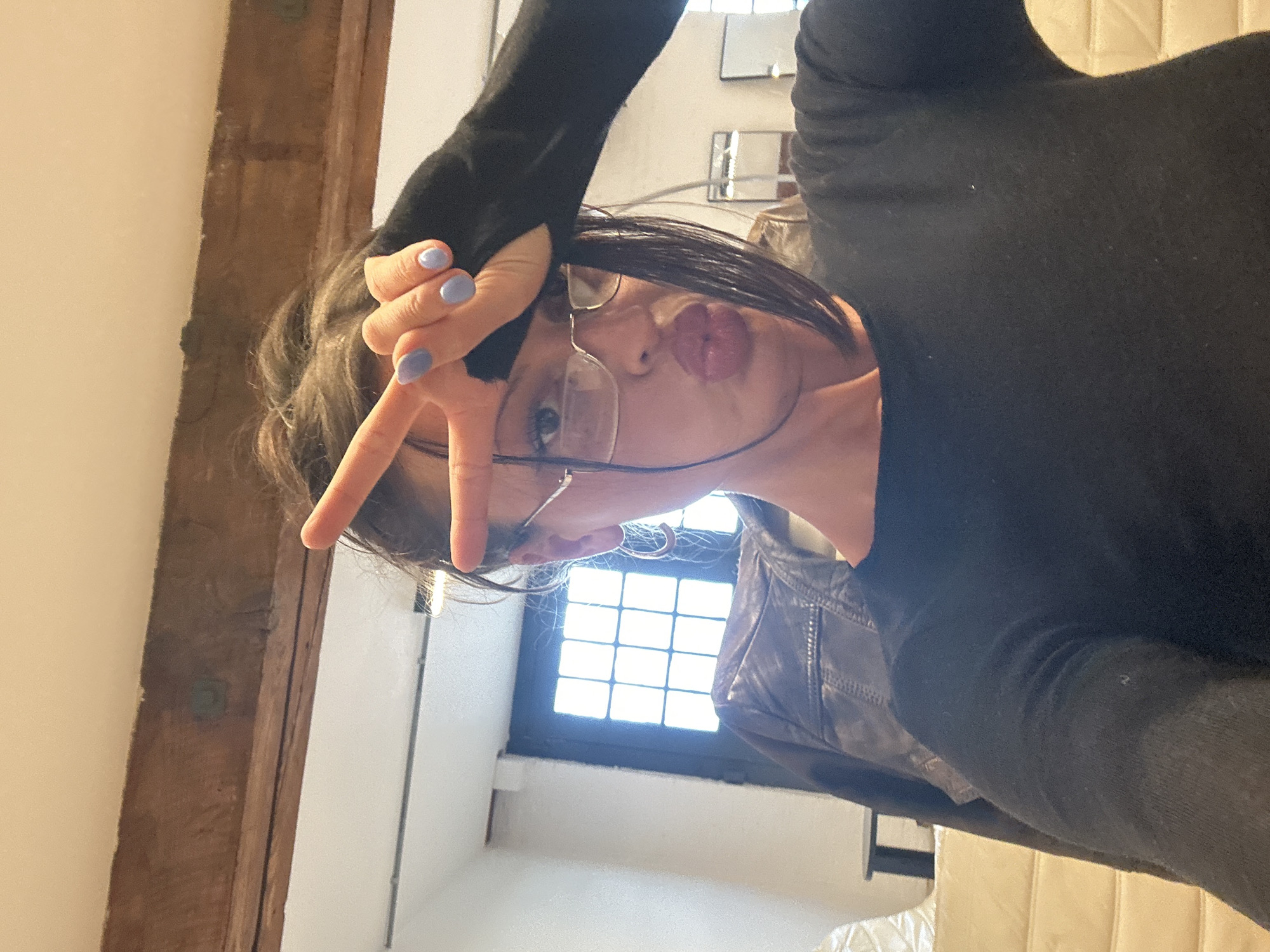 A woman with dark hair, wearing glasses and a black top, is posing indoors. She is making a peace sign with her right hand, positioned near her forehead. Her fingernails are painted light blue. Behind her is a window with multiple panes. To the left, there is a wooden beam and white wall. The woman appears to be wearing a jacket.
It's not possible to determine if this image was taken in the city of London, United Kingdom from the photo.