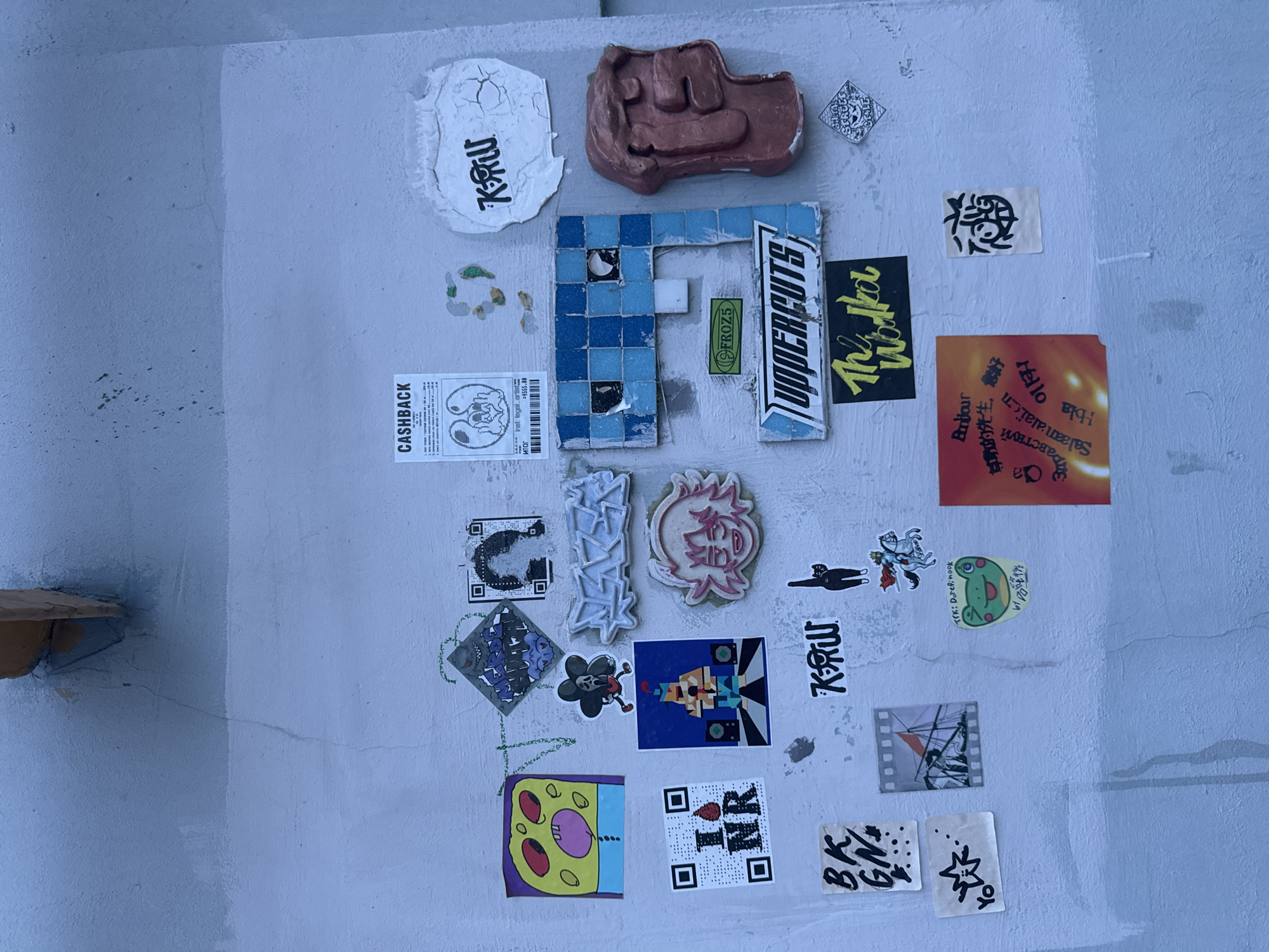 The image shows a variety of stickers and small objects affixed to a light gray, textured wall. There is a mosaic "F" made of blue tiles, a brown fist-shaped object, and numerous stickers with various designs and text, including "CASHBACK," "UPPERCUTS," "FROZ5," "KRW," and QR codes. The text on other stickers is in multiple languages. The wall appears to be part of a building exterior or a wall in a public space. The location of Moscow, Russia is only confirmed by the OCR text and cannot be determined with full certainty based on image contents.