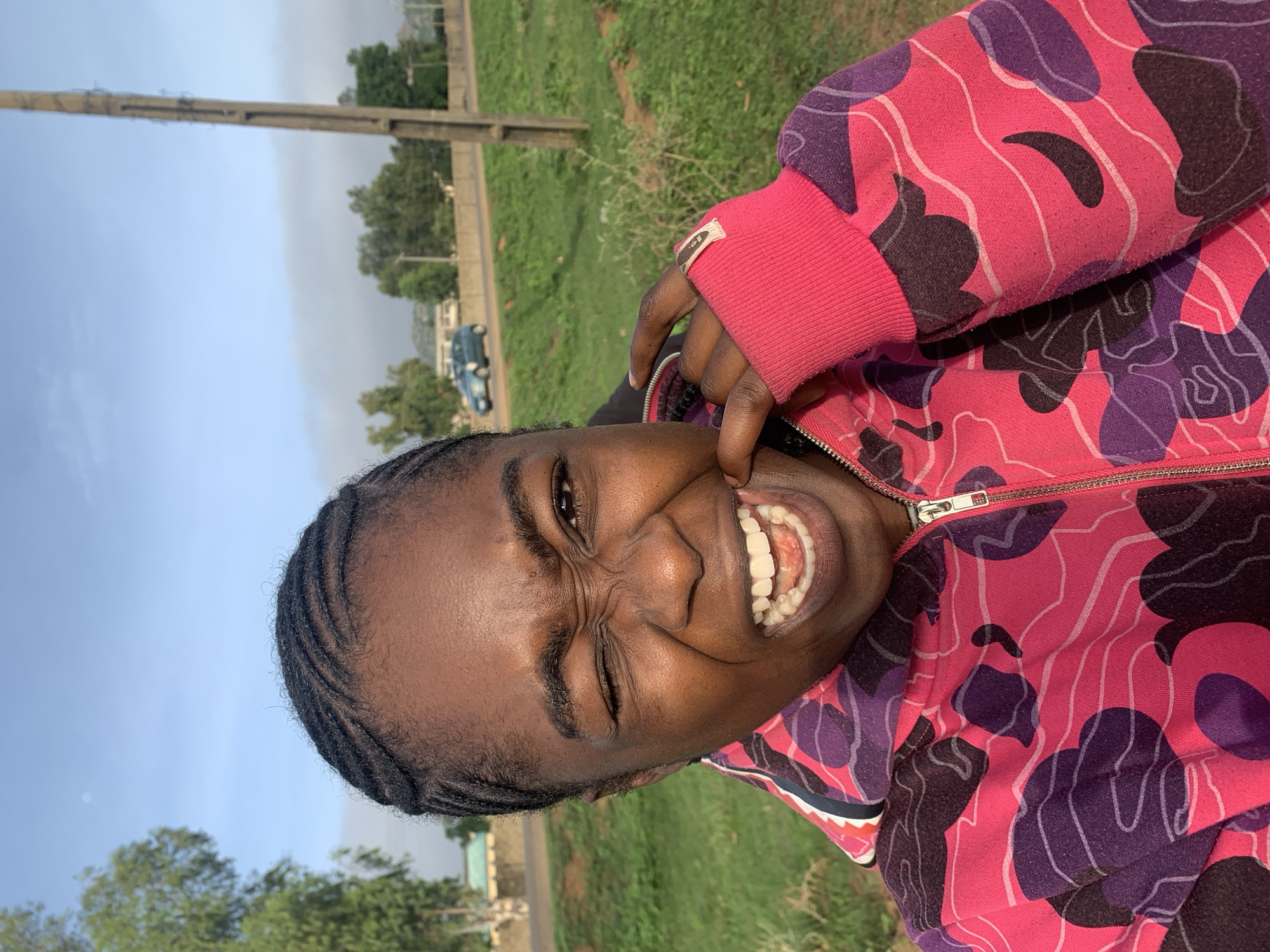 A person with dark skin and braided hair is captured in a close-up, outdoor shot, smiling widely and playfully pulling down their lower lip with their right index finger to expose their teeth and gums. Their eyes are crinkled in a joyful expression. They are wearing a hooded jacket featuring a vibrant, abstract camouflage-like pattern in hot pink, purple, and black, accented with thin white outlines. The jacket has a visible silver zipper.
The setting appears to be a sunny day in an outdoor, somewhat open area. The sky above is a clear light blue, suggesting good weather conditions. In the background, there's a patch of green grass, a dirt road, and several trees with green foliage. A tall utility pole stands vertically on the left side of the frame, and a blue car is partially visible in the distance on the road. The overall atmosphere conveys candid happiness and a relaxed moment outdoors, possibly in Unguwar Saya, Nigeria as indicated by the context. No discernible text is visible on the person's clothing or in the background.