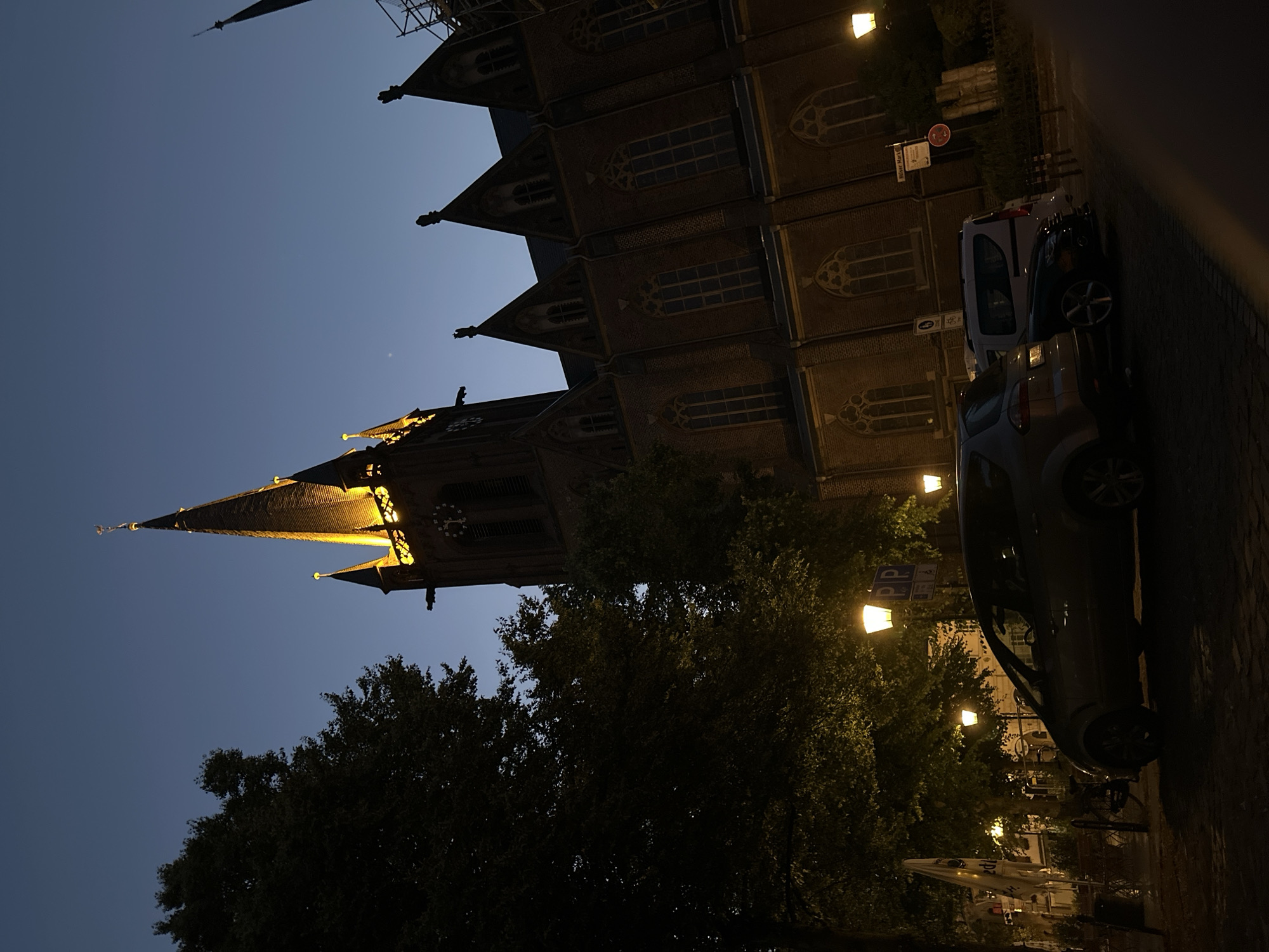 The image depicts a street scene at dusk in Krefeld, Germany, featuring the St. Bonifacius Church. The church, constructed of brick, is illuminated, highlighting its Gothic architecture, pointed spires, and clock tower. In the foreground, several cars are parked on a paved street. Trees with dense foliage line the left side of the image, and streetlights cast a warm glow on the scene. The sky is a deep blue, indicating the transition from daylight to night. No people are visible in the image.