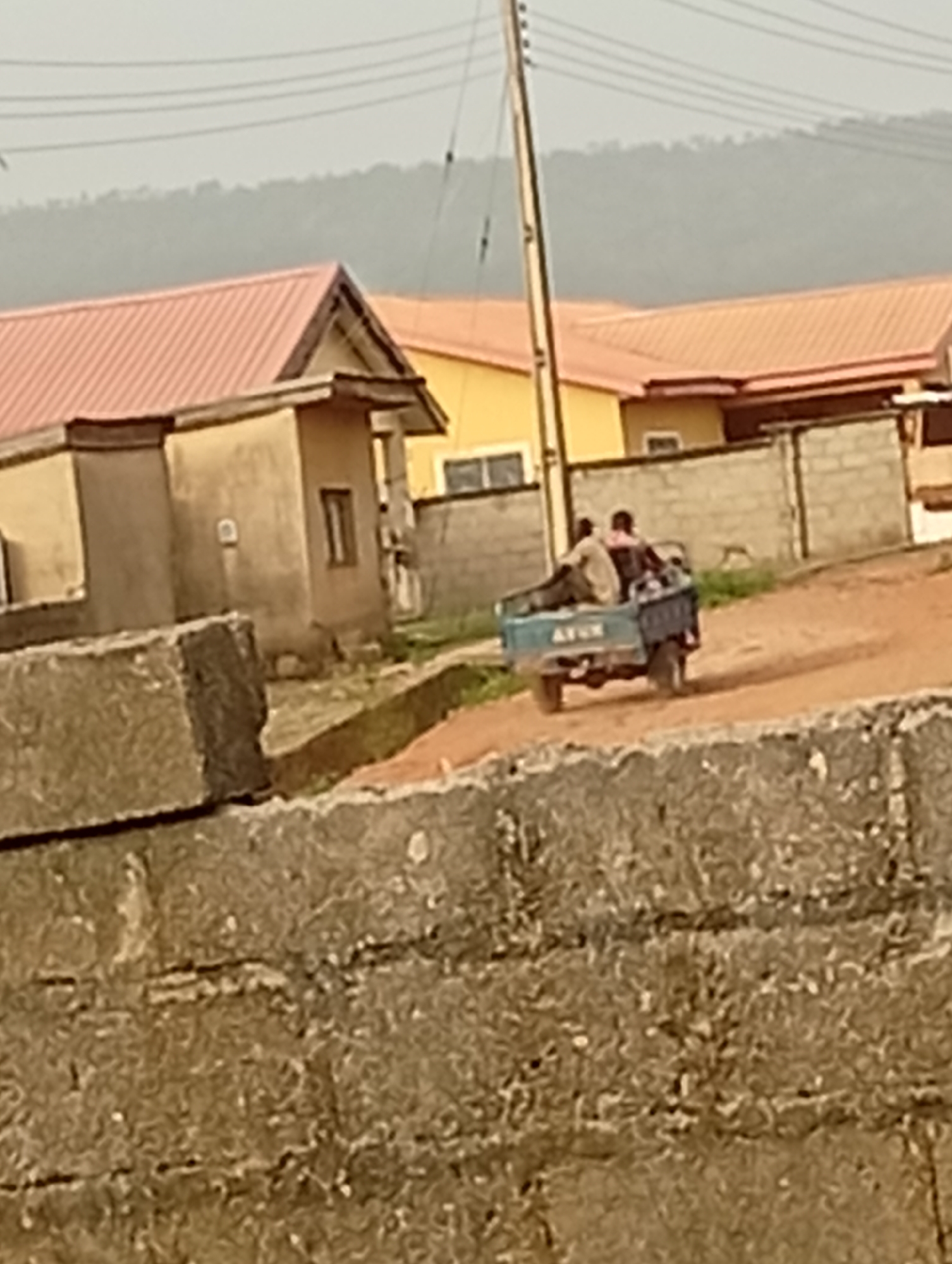 The image depicts a street scene in what appears to be a residential area of Adankolo, Nigeria. In the foreground, a rough, unpainted concrete wall partially obscures the view. Behind the wall, a three-wheeled vehicle with two visible occupants is traveling along a dirt road. The vehicle has lettering on the back, partially readable as "AVON." In the background, there are several single-story buildings with reddish-brown roofs, surrounded by low walls and vegetation. A utility pole and associated power lines are also visible. The overall environment suggests a somewhat rural or less developed part of the city.