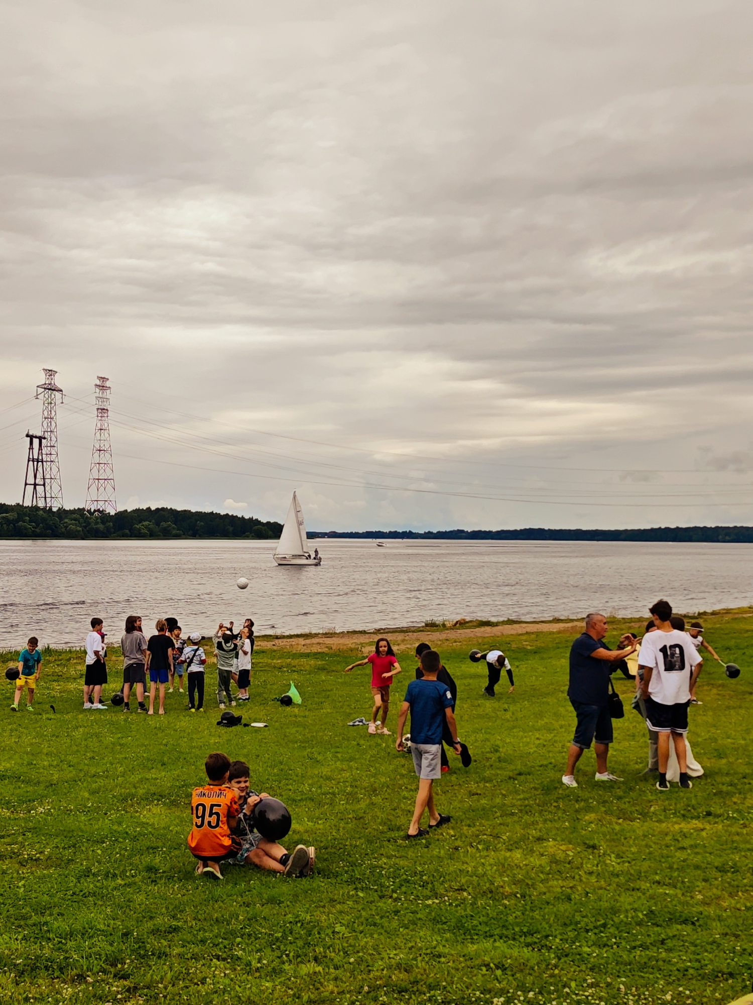 This media file depicts an outdoor gathering near a body of water, likely a river or lake, with a sailboat visible in the distance. The scene is set on a grassy embankment under a cloudy sky, suggesting it is daytime but with overcast weather. In the background, two prominent power line towers stand tall against the horizon, with a dense line of trees bordering the water.
The foreground and midground are populated with a diverse group of people, predominantly children and adolescents, along with a few adults. They appear to be engaged in various leisure activities. Some children are playing with a ball, while others seem to be participating in a game or exercise involving what look like weighted objects. One boy is sitting on the grass with another, holding a black spherical object. An adult male in a blue shirt and shorts is interacting with a young person in a white t-shirt with the number "10" on the back. Another young girl in a red dress is running.
The overall mood seems relaxed and active, indicative of a casual outdoor event or a recreational get-together. The location context of Konakovo, Russia, combined with the presence of a wide body of water and the power line towers, provides specific geographical clues.
Visible text includes the number "95" on an orange jersey worn by one of the boys, and the number "10" on the back of a white t-shirt. The text on the orange jersey also appears to have Cyrillic letters above the number, which might be a name or a team designation.