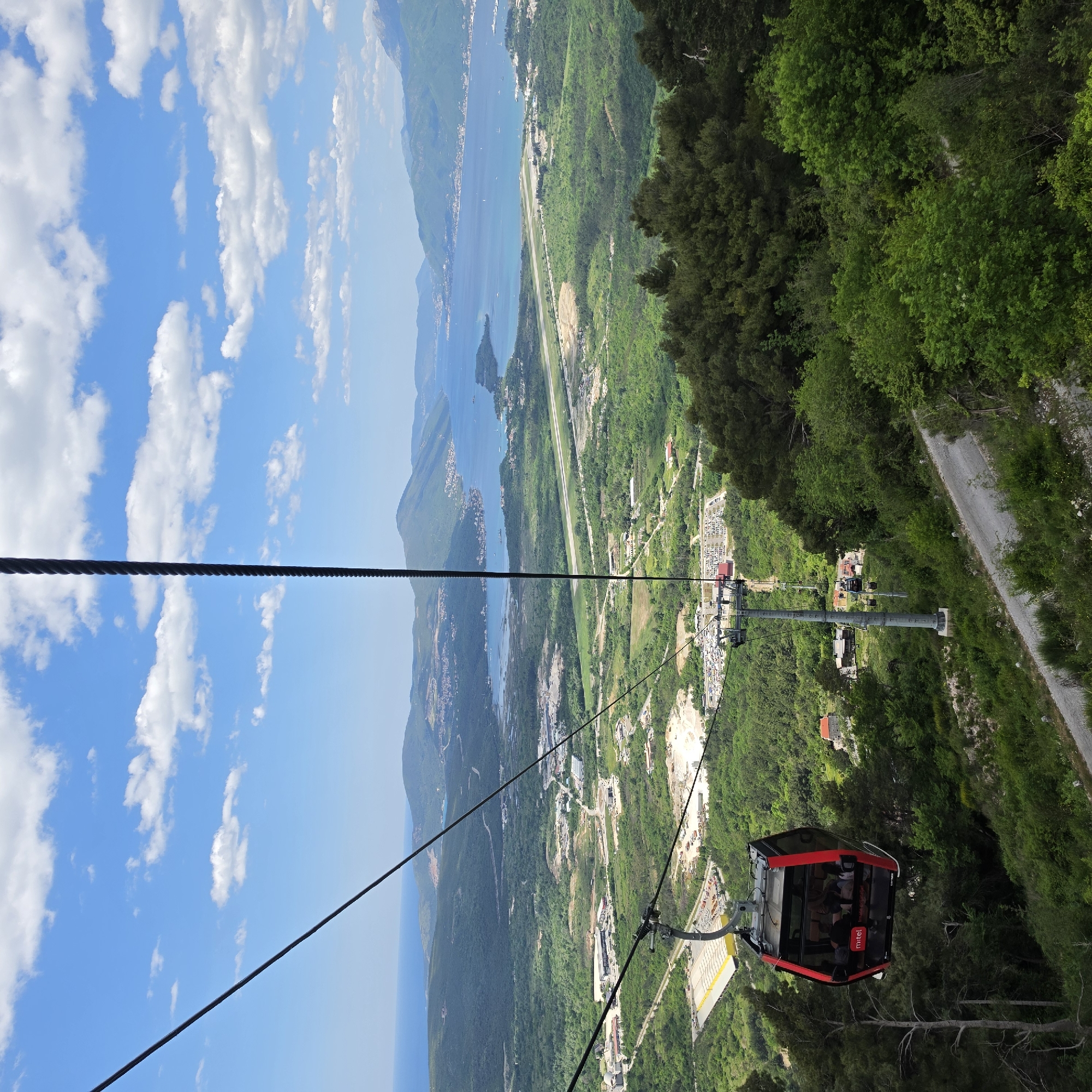 The image shows a cable car descending a steep, forested slope in Čavori, Montenegro. The red and black cable car, labeled "Mt:el," carries visible passengers. The backdrop features a coastal landscape with a clear blue sky dotted with clouds above the Adriatic Sea. The terrain includes green forests, rocky mountains, and a small coastal airport, offering a panoramic view of the bay. The cable car cables are visible extending from the top left of the image to the car.
