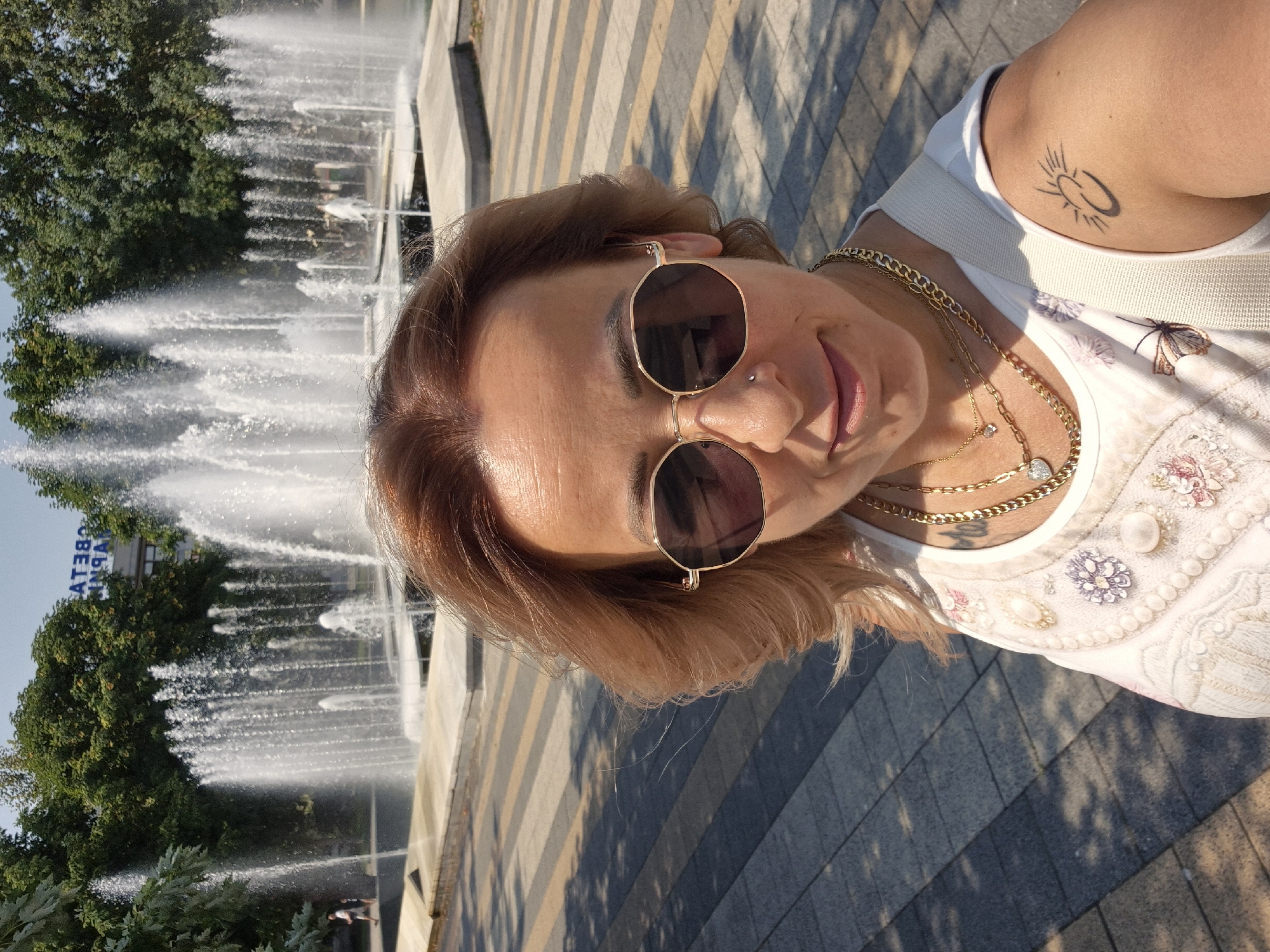 In the city of Unknown, Unknown, a woman with shoulder-length auburn hair, wearing gold-rimmed hexagonal sunglasses, a white top adorned with floral embroidery and pearls, and multiple gold necklaces, takes a selfie in front of a multi-tiered fountain. A small sun tattoo is visible on her upper right arm. The fountain is active, with numerous water jets creating a misty spray. The ground is paved with a pattern of grey and yellow rectangular tiles, with shadows cast by unseen trees. In the background, lush green trees line the top of the frame, and a building with text, partially obscured, is visible above the trees. A few individuals are faintly visible walking in the distance near the fountain. The overall scene suggests a public outdoor space on a sunny day.