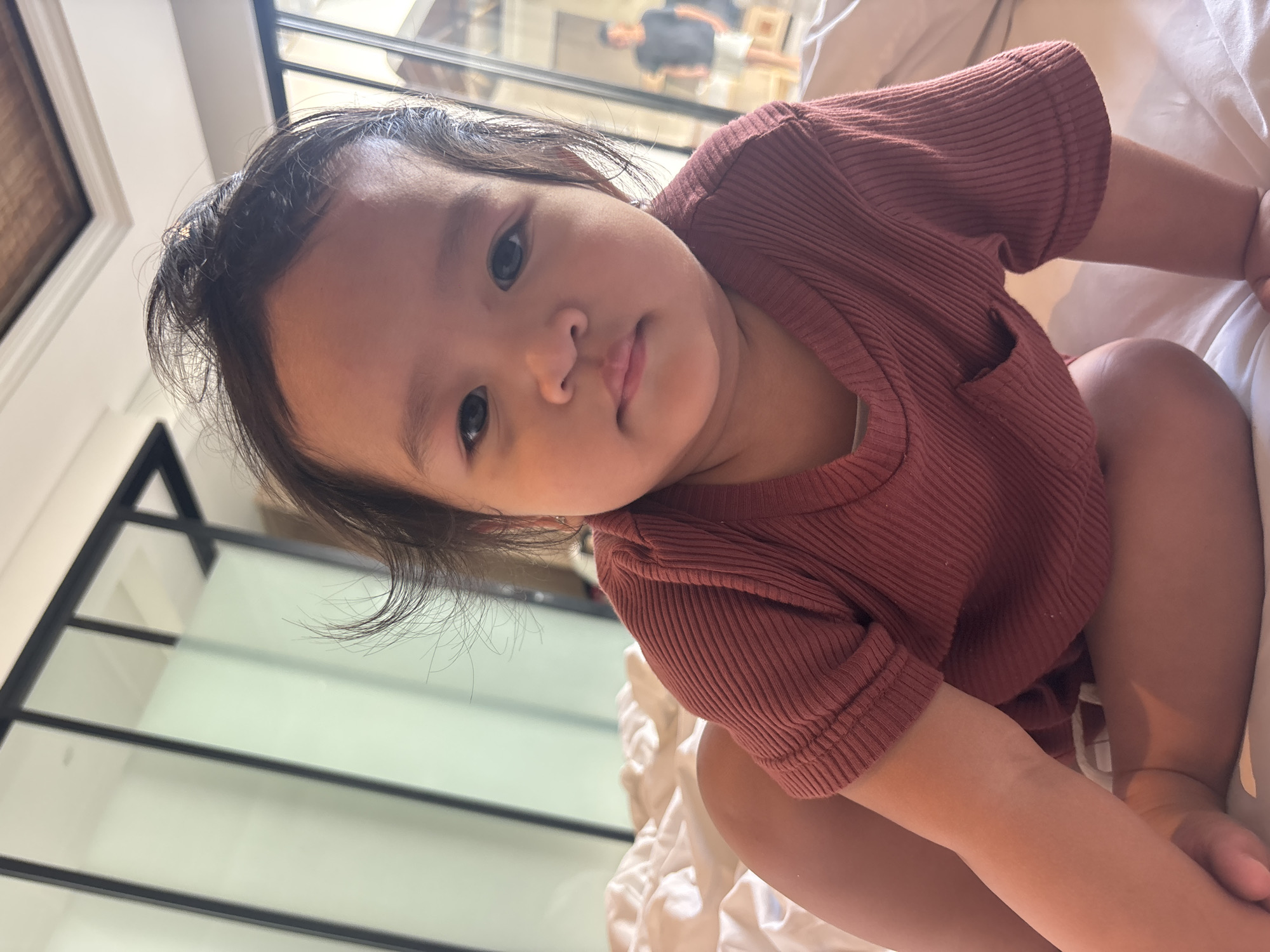 A young child with dark hair and dark eyes is sitting on a bed. The child is wearing a ribbed, maroon-colored t-shirt. The child's hair is messy and some strands are sticking up. In the background, there is a glass partition with black frames. Through the glass, another person is visible in the distance, wearing a dark t-shirt and shorts. The room appears to be a bedroom, possibly in a hotel or resort in North Kuta, Indonesia, suggested by the interior design elements. The bed is covered with white bedding.