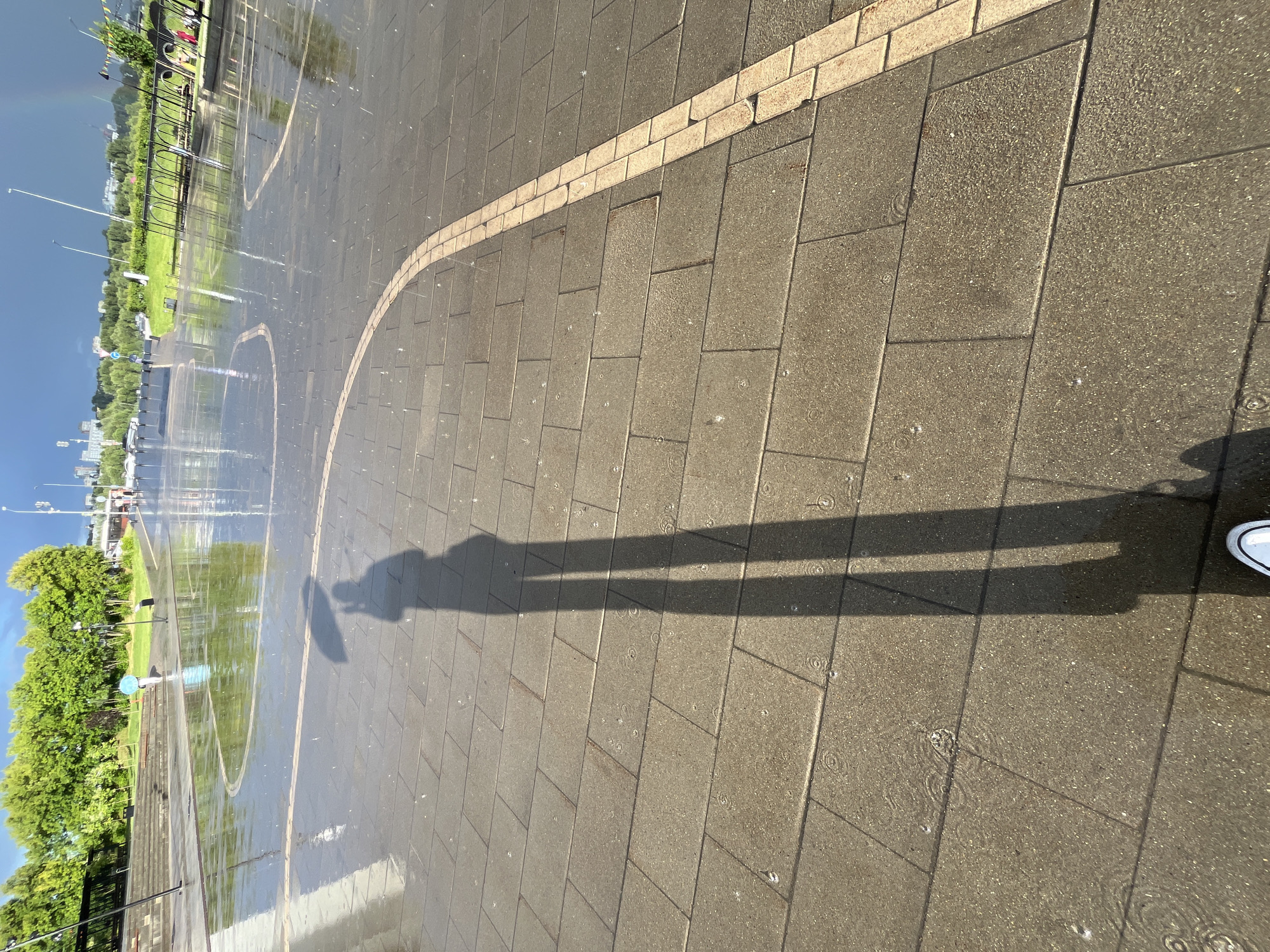 The image depicts an outdoor scene with a wet, paved area in the foreground, suggesting recent rain. The paving stones are dark gray, interspersed with a lighter, cream-colored border that curves across the frame. Small ripples and scattered droplets are visible on the wet surface, indicating ongoing light rain or residual moisture. A prominent shadow of a person holding an umbrella stretches across the paving stones, its shape suggesting the person is standing to the left of the frame. One white shoe with black laces is visible at the bottom right corner of the image, also casting a shadow.
In the background, a green lawn is visible, which is also wet and reflective. Circular markings, possibly for a game or fountain area, are faintly discernible on the wet grass and pavement. A row of lush green trees lines the edge of the lawn. Beyond the trees, a dark, moody sky dominates, with a hint of a rainbow arcing on the left side. In the distance, urban elements, including buildings and lampposts, are visible, suggesting a park or public space within a city. The overall lighting indicates it is daytime, likely late afternoon or early evening, given the long shadows. The presence of rain, the umbrella, and the wet surfaces point to a rainy or recently rainy weather condition. The scene evokes a sense of dampness and perhaps a fleeting moment of beauty with the rainbow. There is no visible text in the image.