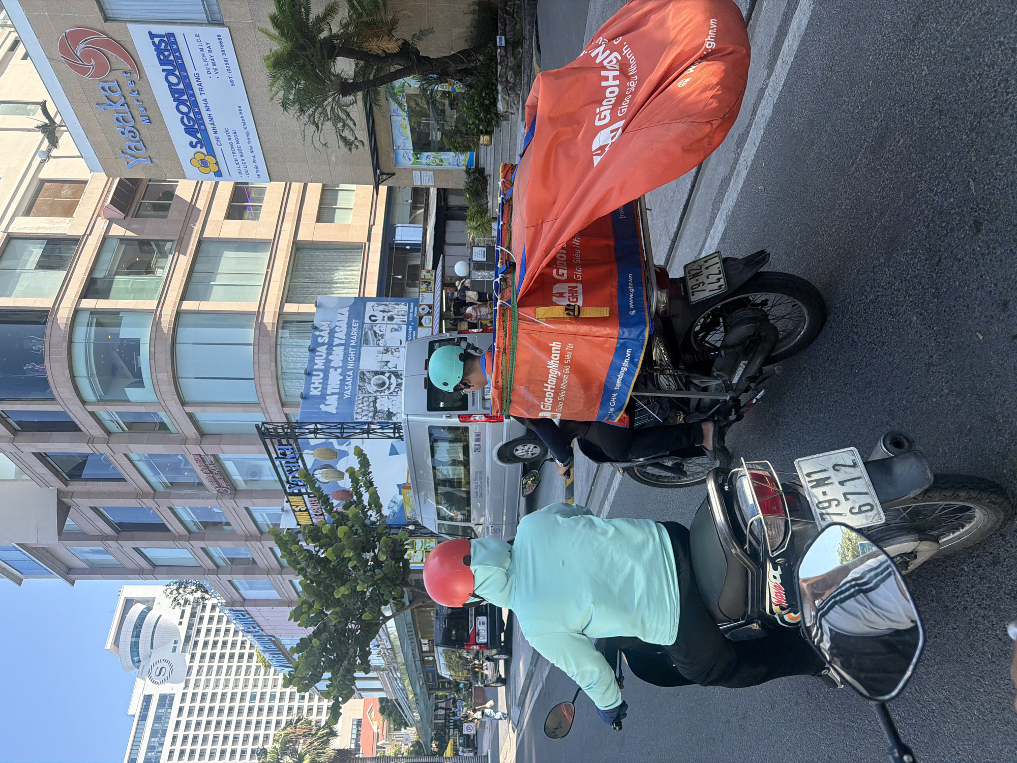 Two individuals are riding motorcycles on a city street. The motorcycle in the foreground is ridden by a person wearing a light blue hooded jacket and a red helmet. The motorcycle in the background is ridden by a person wearing a teal helmet and a dark top, with a large orange bag strapped to the back of their motorcycle, labeled with "GiaoHangNhanh". The motorcycles are on a paved road with a white lane marker. In the background, there are multi-story buildings, including a tall white building with a distinct curved top, and a building with "Yasaka Market" signage. Palm trees are visible on the right side of the image. The scene appears to be in Nha Trang Ward, Vietnam, as indicated by the building signage.