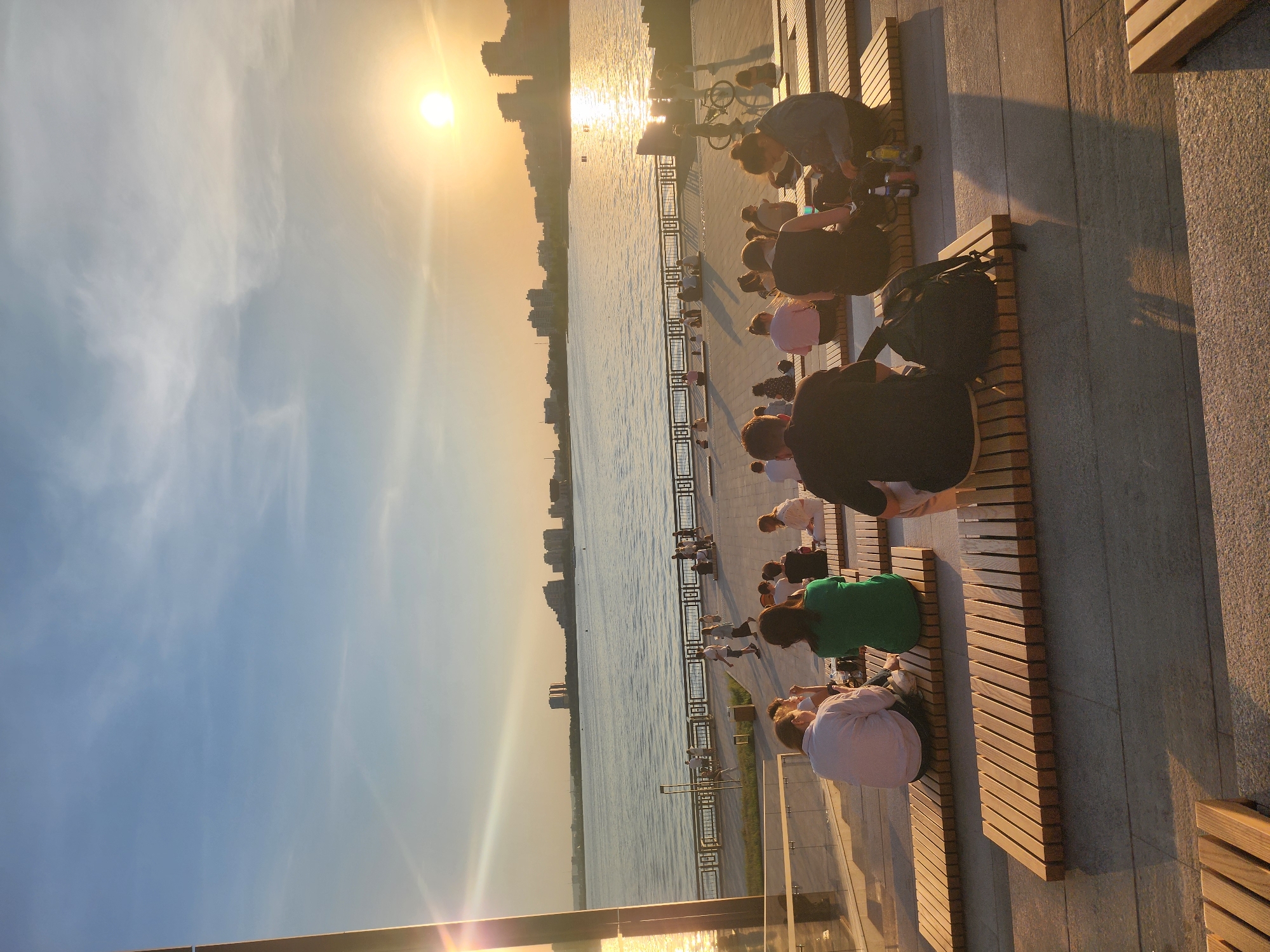 The image captures a sunny, likely summer, scene on a waterfront promenade in Kazan, Russia. The promenade, likely the Kazan Kremlin Embankment, is lined with wooden benches where people are sitting and engaging in various activities. Some individuals appear to be resting, while others seem to be socializing or observing the environment. There is a river or large body of water in the background reflecting the bright sunlight, with a city skyline visible on the opposite bank. The sky is clear with some wispy clouds. The overall ambiance suggests a relaxed and pleasant atmosphere in a public space. There is a bicycle in the background and various other people walking, suggesting pedestrian activity.