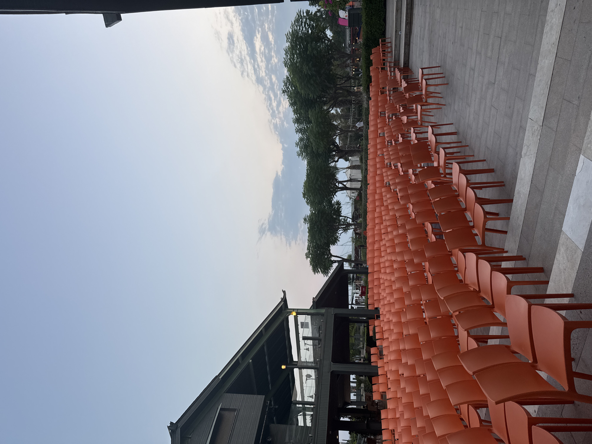 An outdoor amphitheater seating area is shown in Aksu, Turkey, during twilight. Rows of orange plastic chairs stretch across a paved area, facing a stage area not fully visible. To the left, a dark, multi-level building with a sloped roof and open balconies is present. Trees line the background, some with lush green foliage. The sky is a pale blue with scattered clouds, indicating the transition from day to night. No people are actively engaged in any activity, and the scene appears set for an event.