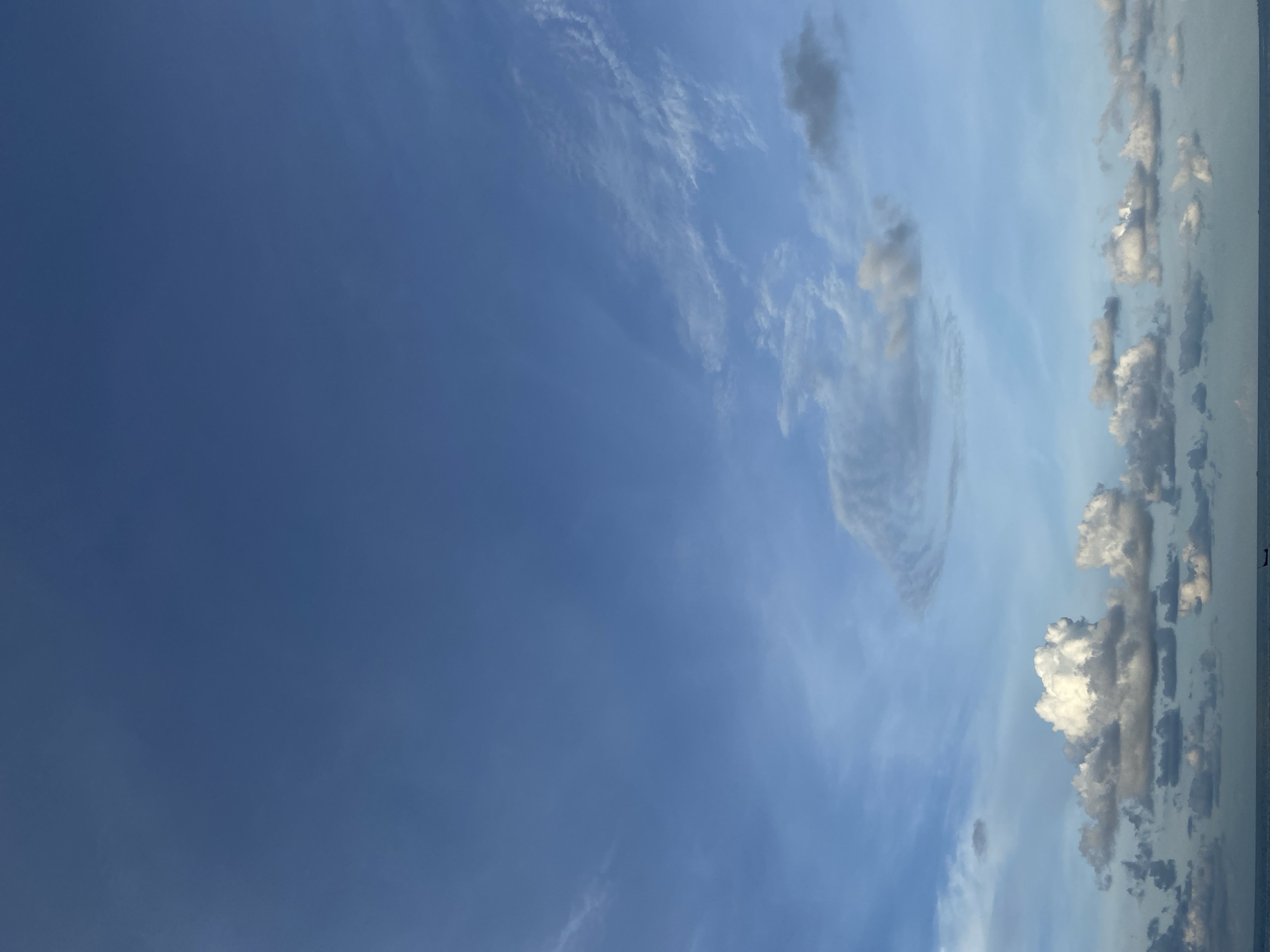The image presents a daytime sky in Lhokseumawe, Indonesia. The sky transitions from deep blue on the left to a lighter blue with a variety of clouds towards the right. A distinct, circular cloud formation is visible, and numerous puffy cumulus clouds are present near the horizon on the right. The horizon itself is faintly visible. No people or man-made objects other than the clouds are apparent in the frame.
