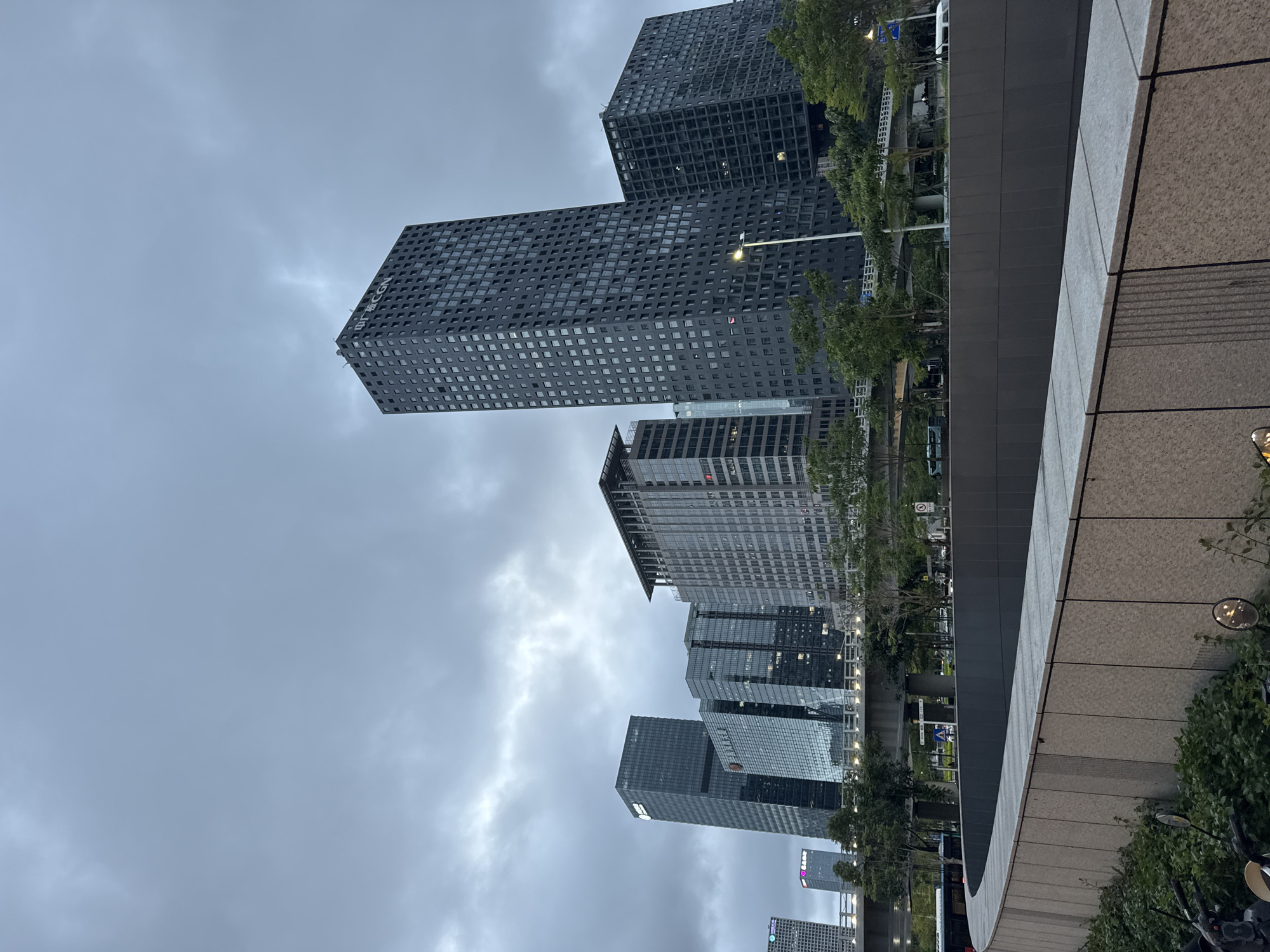 The image captures a low-angle view of several skyscrapers in the Futian District, China. The sky is overcast with gray clouds. The buildings are primarily modern, with glass and steel facades. A large, dark building with a grid-like pattern is prominently visible. In the foreground, a stone or concrete wall extends across the lower right of the frame. Green trees are interspersed among the buildings along the street. The photograph appears to have been taken from ground level.