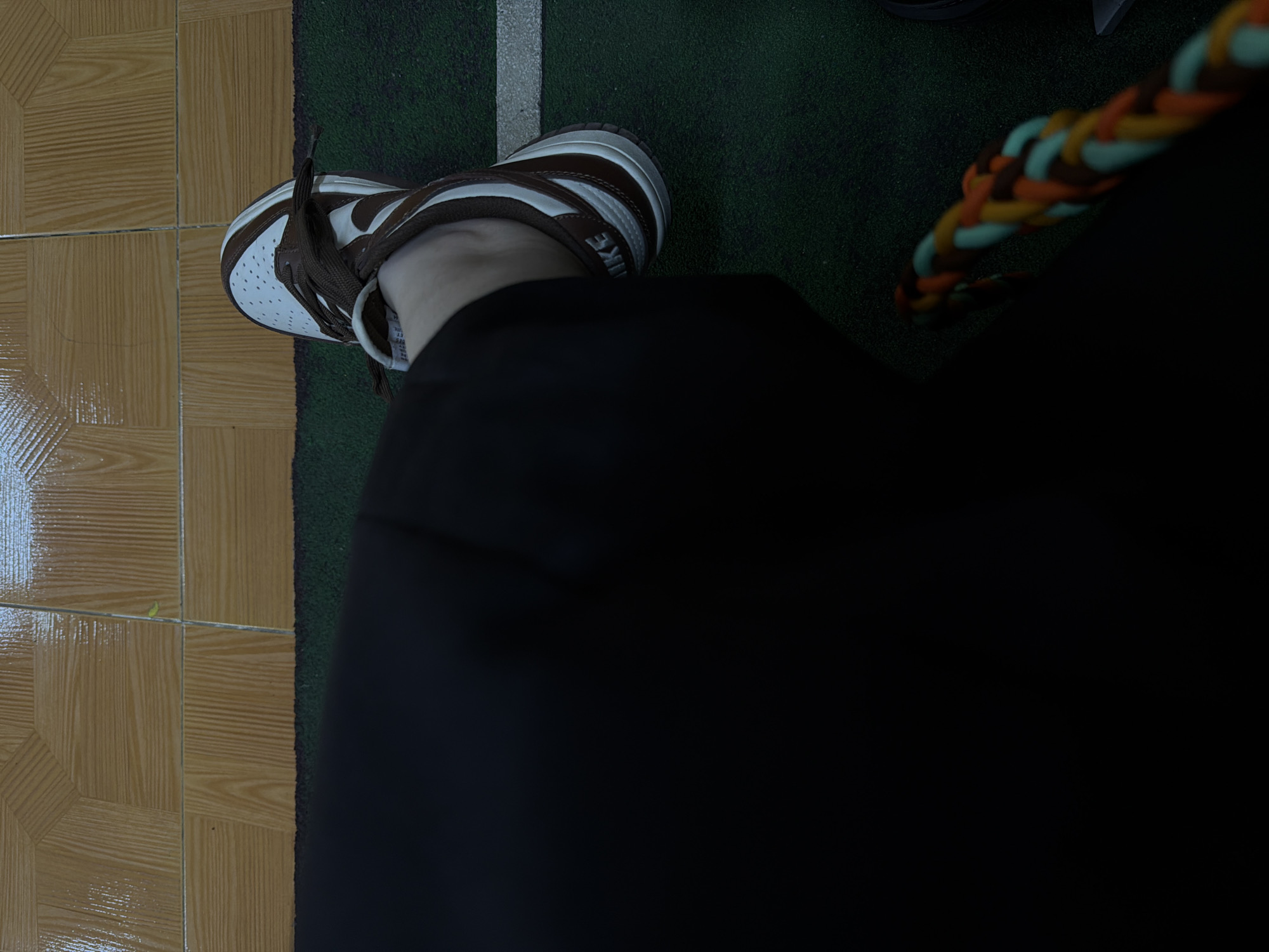 A top-down shot captures a single brown and white Nike Dunk sneaker, primarily situated on a dark green artificial turf surface marked with a white line. A small section of brown tiled flooring is visible on the left edge. The sneaker is worn by an individual dressed in black pants, with the lower portion of their left leg visible. The context of the image suggests an indoor or semi-indoor sports facility. There is no explicit reference to the city of Yerevan, Armenia.