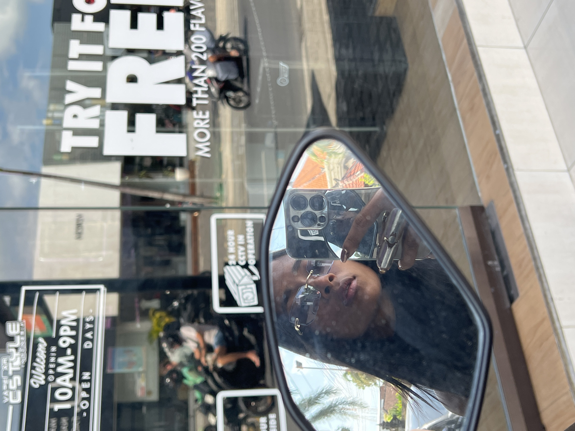 A young woman with long dark hair is taking a selfie in a motorcycle mirror. She is wearing sunglasses and a dark top. In the background, through a glass window, there is a street scene in North Kuta, Indonesia. Signs on the window advertise "VAPE & JUICE INDONESIA" and "TRY IT FOR FREE". There are motorcycles with riders on the street, and palm trees are visible. The overall lighting suggests it is daytime.