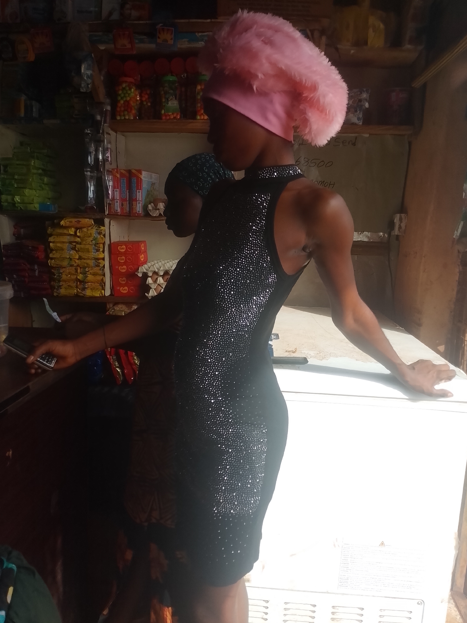 In Ejule, Nigeria, a young woman stands in a small shop or convenience store. She wears a black, sleeveless, knee-length dress covered in sparkling embellishments and a large, pink, fuzzy head covering. Another person, a child, stands behind her. The woman rests her left hand on a dark counter near a cash register or point-of-sale system, and her right hand rests on the top of a bright, white freezer. The shop is stocked with various food items and household goods on shelves behind her. A price list is written on the wall.