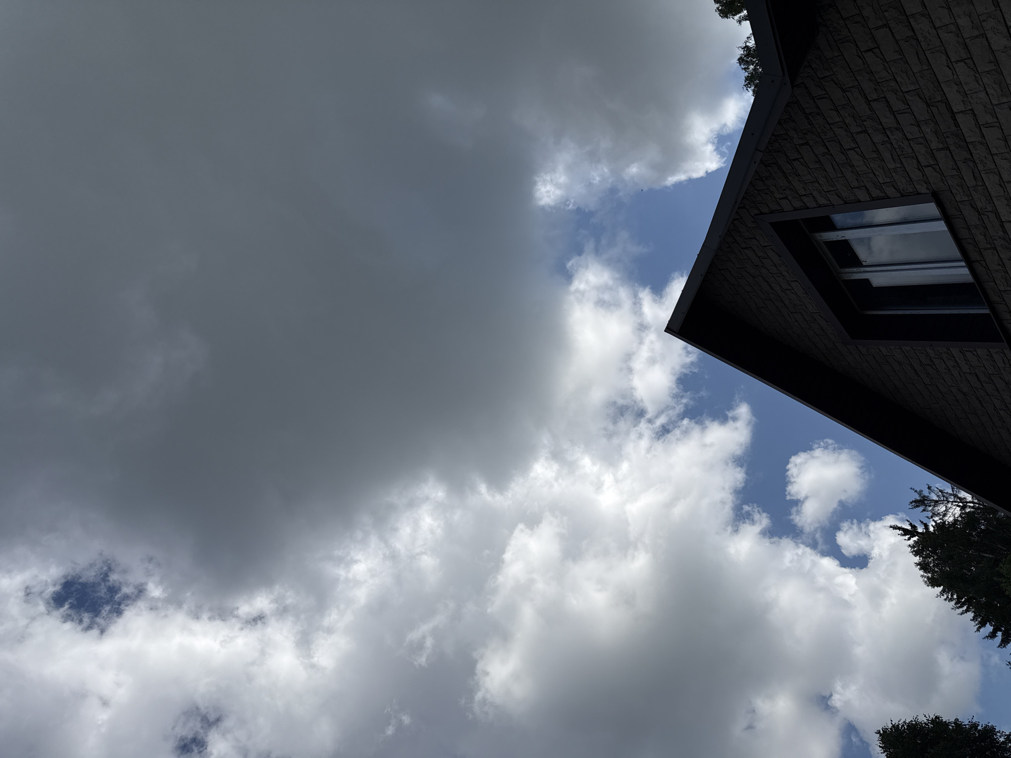 The image shows a close-up, low-angle view of the roof and upper wall of a brick house under a cloudy sky. The brickwork is a dark brown color with a rough texture. A single window with reflective panes is visible on the upper wall. The roof is dark gray and steeply pitched. In the background, the sky is filled with a mix of bright white, fluffy clouds and darker, more ominous gray clouds. Patches of blue sky are visible between the clouds. The tops of some dark green evergreen trees are visible along the bottom edge of the frame, to the left and right of the house. The scene appears to be in a residential area. The image was taken in Mahilyow, Belarus.