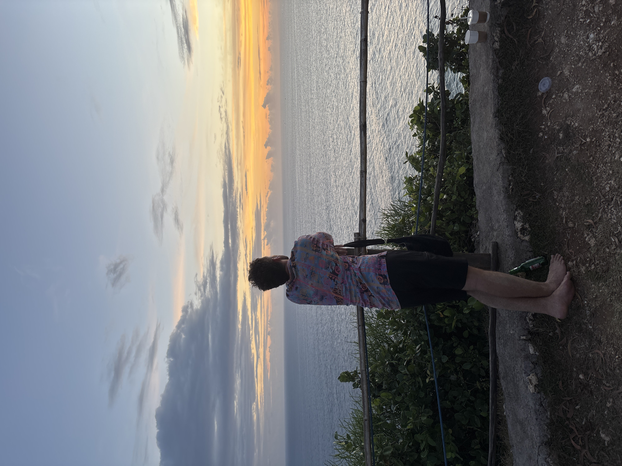 A man stands barefoot on a ledge overlooking the ocean in Kuta Selatan, Indonesia. He wears a colorful shirt and black shorts. He rests his arms on a bamboo railing. Behind the railing are green plants, while beyond it is the ocean and a sunset sky with orange and grey clouds. Discarded coffee cups are visible to the right. The scene appears casual. An empty green bottle is by his feet.
