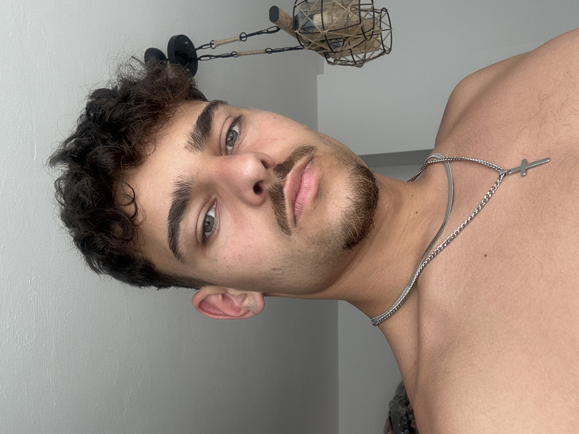 A close-up, vertically-oriented image shows a fair-skinned male with curly dark hair, prominent eyebrows, and green eyes, looking directly at the camera. He has a trimmed mustache and beard. The man is wearing a silver necklace with a cross pendant. He is partially undressed, revealing his chest and upper shoulder. The background is a light grey wall. At the top of the image, a decorative light fixture with a woven shade is visible. There is no information to indicate that this image was taken in the city of Troyan, Bulgaria.