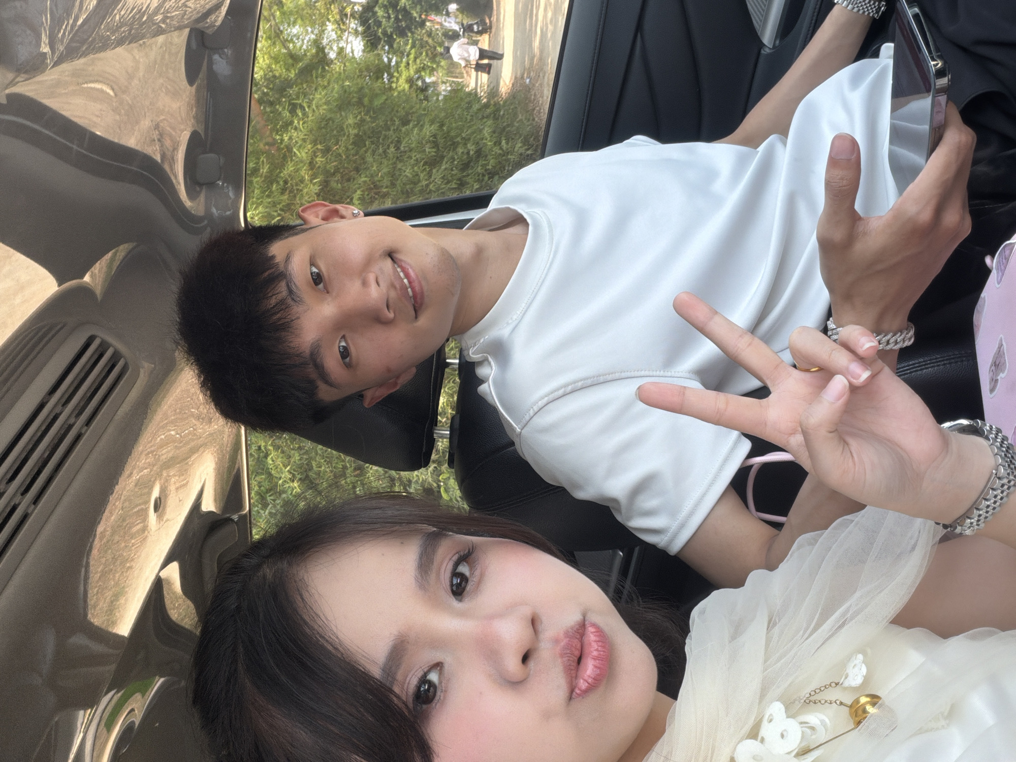 A selfie taken inside a car captures two young adults, a man and a woman, posing for the camera. The image is oriented vertically, showing them from an overhead perspective.
The man, positioned further back and to the right (when viewing the image upright), wears a white t-shirt and has short dark hair. He smiles warmly at the camera, holding a modern smartphone in his left hand, and making a peace sign with his right hand. He wears a small earring in his left ear and a silver-toned chain bracelet on his right wrist.
Closer to the camera and to the left, the woman has long dark hair and wears a light-colored top, possibly white or cream, featuring a sheer, delicate overlay. Her face shows makeup, including blush and lip gloss, and she has a gently pouting expression for the photo. A silver-toned watch adorns her left wrist. On her top, a decorative gold-colored brooch with white, possibly floral or butterfly-shaped, accents is visible. Her right hand also appears to make a peace sign gesture, positioned near the man's hands.
The setting is the interior of a vehicle, with parts of the car's dashboard, air vents, and a door panel visible. Through the car window in the background, green foliage, trees, and what appears to be an outdoor path or road are discernible. Two distant figures, including one in a white shirt, can be seen walking. The natural light suggests it is daytime, possibly sunny. The man's smile conveys a cheerful mood. The scene takes place in Bình An, Vietnam. There is no visible text in the image.