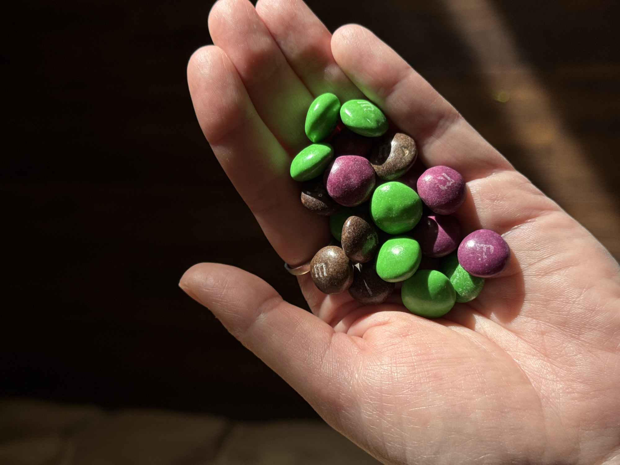 A hand holds a collection of green, purple, and brown M&M candies against a dark background. The scene is illuminated by a light source from the left, casting shadows across the hand and candies. No information indicates a location in the city of Chaikovsky, Russia.