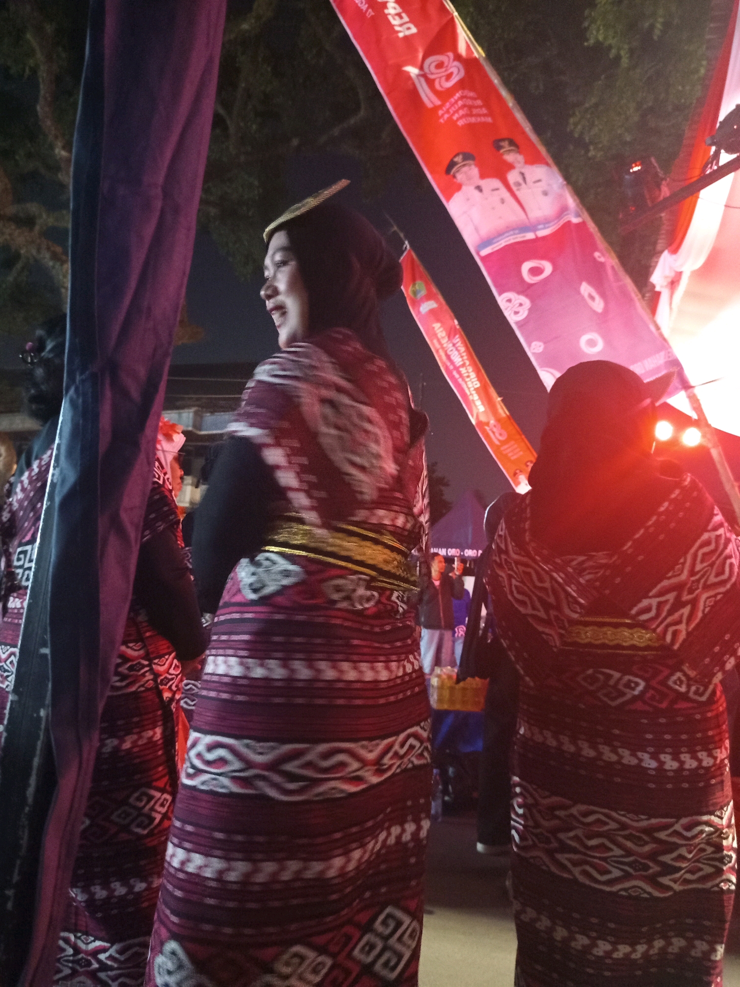 The image captures a scene at night, likely during a festival or event, featuring individuals in traditional attire. In the foreground, a person is seen from the back, wearing a dark headscarf and a richly patterned red and white garment. This garment appears to be a sarong or a long skirt with intricate geometric designs. To the left, another individual in similar traditional clothing is partially visible, with a purple fabric also obscuring part of the frame. In the background, red and white banners with text and images of two men in uniform are displayed. The lighting suggests an outdoor setting, with the glow of lights visible in the distance and shadows cast by the surrounding trees. There are no explicit indicators in the image to pinpoint the exact location within Malang, Indonesia, but the general atmosphere suggests a public gathering.