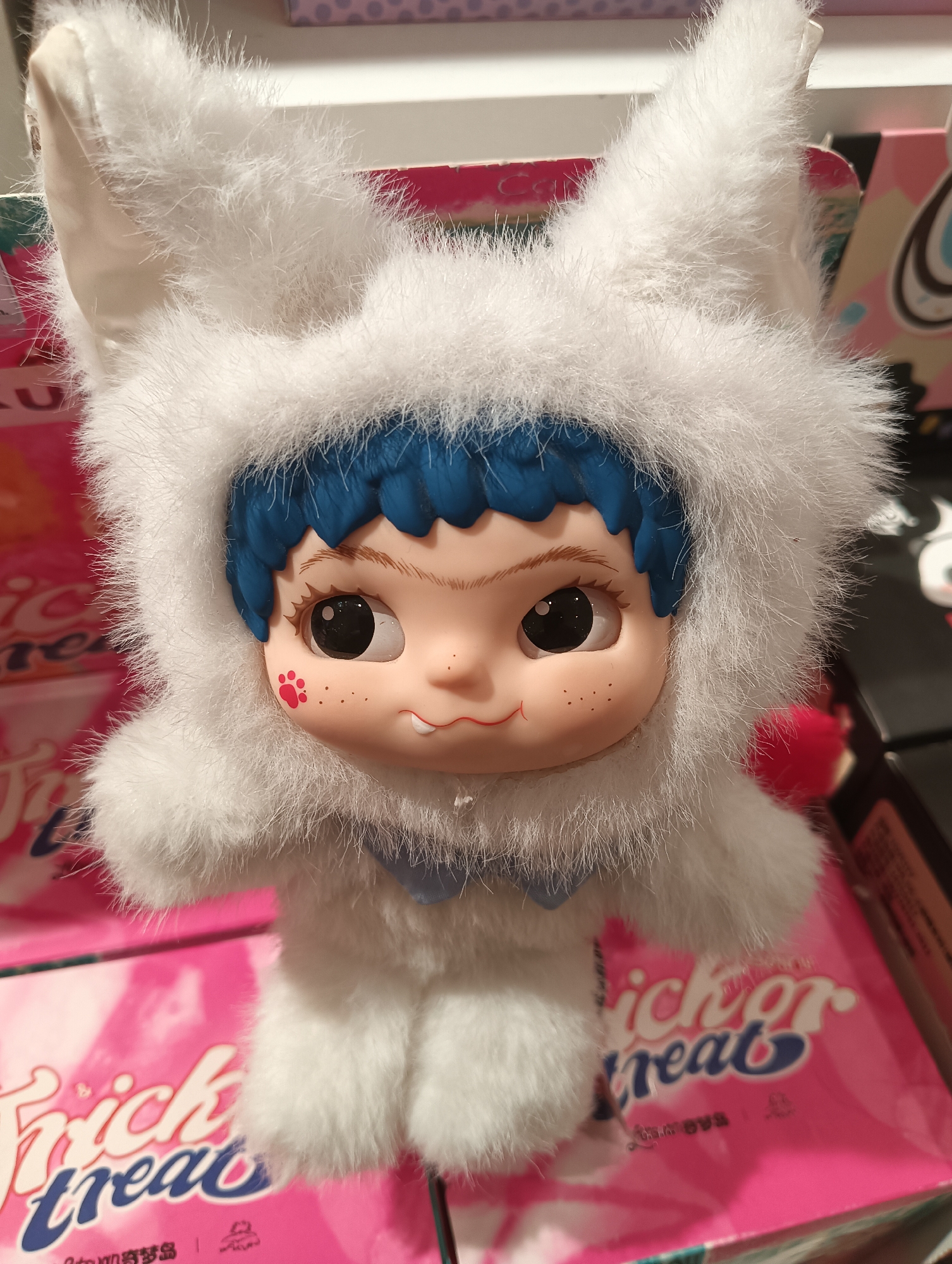 The image shows a close-up, eye-level shot of a doll with blue hair and a fluffy white hooded costume resembling a rabbit. The doll has large, expressive eyes, a small mouth with visible teeth, and freckles on its cheeks, with a red paw print on its left cheek. The doll is positioned in front of pink boxes with the text "Trick or Treat" and other Chinese characters printed on them. The background is blurred, suggesting it is a retail environment. The lighting is bright and diffused. No specific location in Kediri City, Indonesia can be determined from the image.