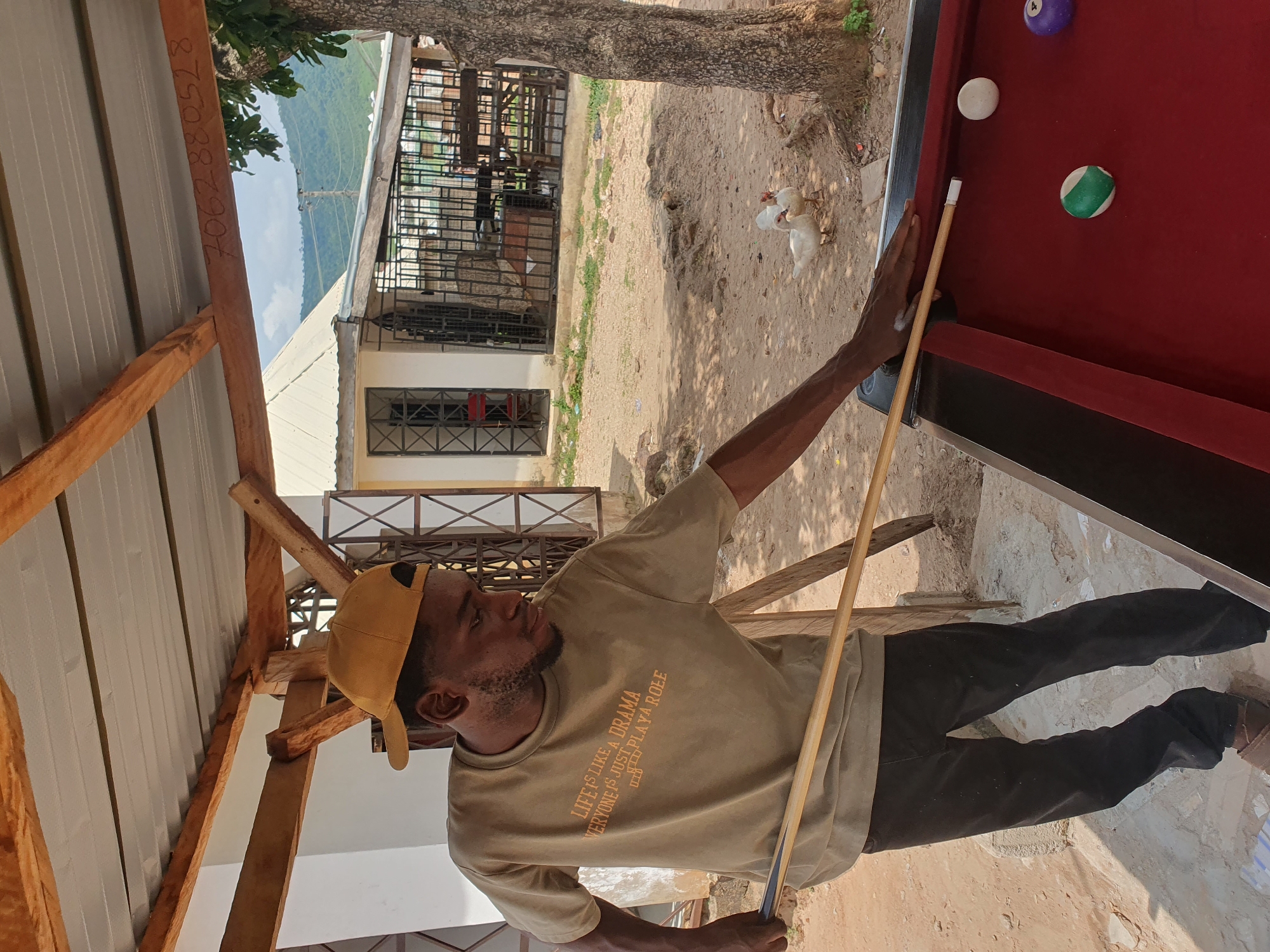 The image shows a young Black man wearing a tan t-shirt and black pants, with a yellow baseball cap, playing pool outdoors. He is holding a cue stick and aiming at the balls on the red felt table. The t-shirt has text printed on it: "LIFE IS LIKE A DRAMA EVERYONE IS JUST PLAYA ROLE". The scene is set in Lokoja, Nigeria, under a covered outdoor area with a corrugated metal roof supported by wooden beams. In the background, there is a white building with barred windows, a fence, and trees. Two white ducks are visible on the sandy ground to the right of the pool table. The overall environment suggests a casual, rural, or semi-urban setting.