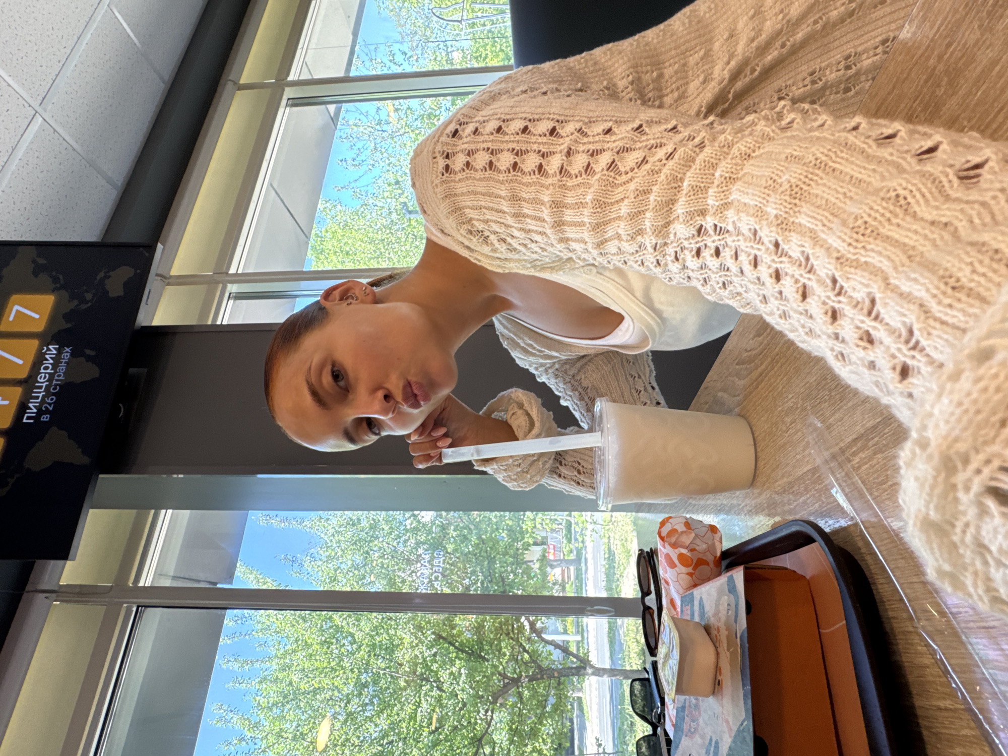The image is an indoor selfie taken by a woman inside a McDonald's restaurant. She is seated at a wooden table near a large window that overlooks a street with trees and green foliage. She is wearing a cream-colored knit sweater and a white top. On the table is a McDonald's tray with a drink in a white cup, a McDonald's container, and a pair of sunglasses. A television screen is visible in the background. Based on the signage in Russian language visible on the screen, the location is identified as Chaikovsky, Russia. The woman is holding a straw and making a kissing face for the camera.