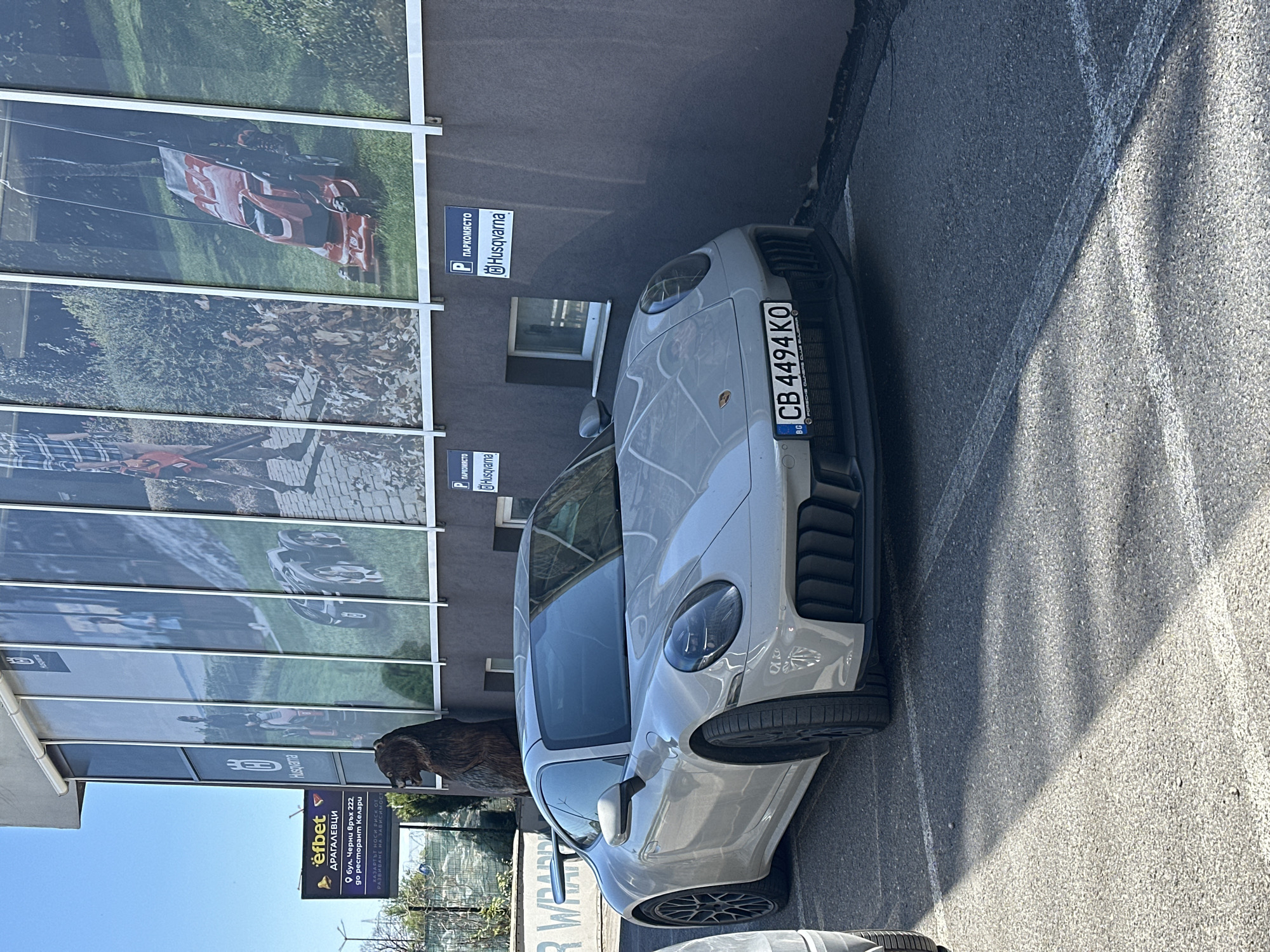 The image shows a light gray Porsche parked next to a building with advertisements for Husqvarna. The license plate of the car is "CB 4494 KO" which indicates it is registered in Sofia, Bulgaria. The area is likely a commercial zone as indicated by the business signs and parking spaces. A sign for "efbet ДРАГАЛЕВЦИ" is also visible, indicating the location is in the Dragalevtsi neighborhood of Sofia, Bulgaria, specifically on Cherni Vrah Boulevard 222. A wooden bear statue stands next to the Porsche.