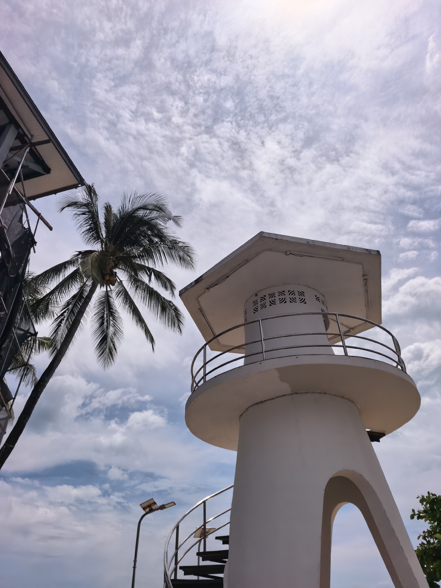 The image depicts a low-angle, wide shot of a white, multi-tiered cylindrical tower with a platform and railings, possibly a lookout or a small lighthouse, situated outdoors in a tropical environment. A tall, slender palm tree with coconuts is to the left of the tower, its fronds reaching towards the sky. To the far left, a portion of a modern building with a slanted roof and exposed metal structure is visible. In the foreground, a spiral staircase made of dark metal, with railings, leads upwards. A street lamp is positioned on a pole to the left of the staircase. The sky above is filled with scattered, wispy white clouds against a pale blue backdrop, with a bright area suggesting sunlight filtering through. The overall scene suggests a coastal or elevated location, possibly within the district of Ko Samui, Thailand, characterized by its tropical vegetation and modern infrastructure.
