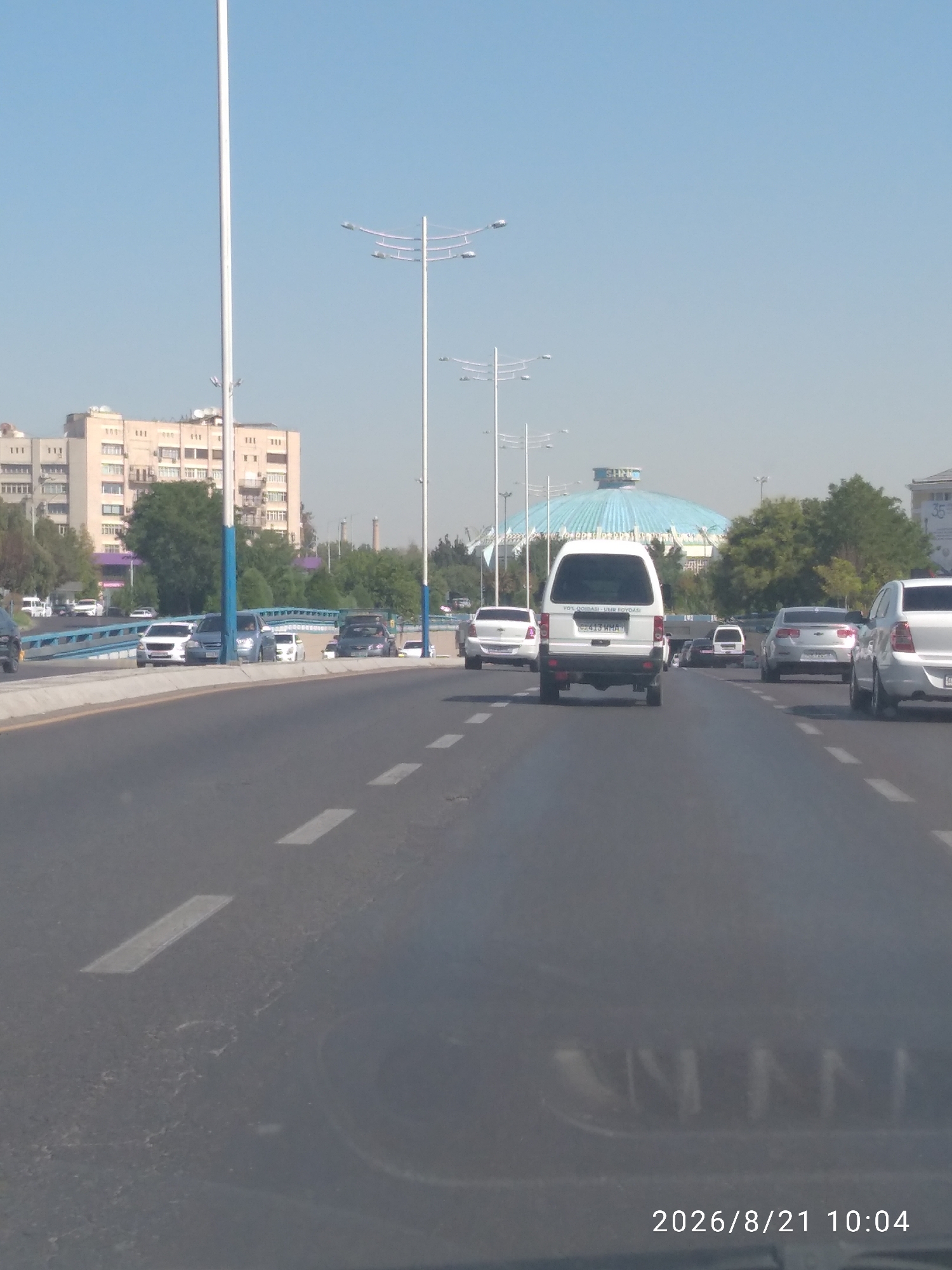 The image depicts a street scene in Tashkent, Uzbekistan, on a clear day. The perspective is from inside a moving vehicle, looking forward. The road has lane markings and appears to be a multi-lane thoroughfare. Several vehicles are visible on the road, including white sedans, a white SUV, and a dark-colored sedan.
In the background, a light blue domed structure, possibly a circus or entertainment venue, is prominent. To the left of this dome, a tall apartment building with multiple windows and balconies can be seen. Several lampposts line the street, characteristic of urban infrastructure. Trees and other foliage are present, indicating a green urban environment. The sky is a clear, pale blue, suggesting fair weather. A timestamp in the lower right corner reads "2026/8/21 10:04".