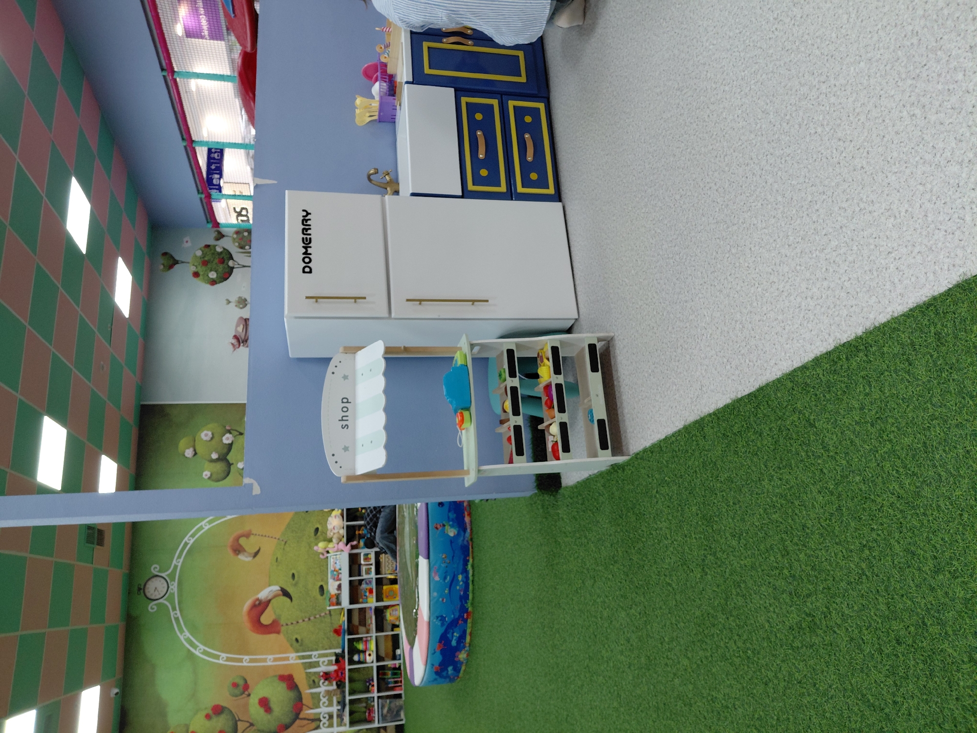 This is a full-shot photograph of an indoor play area, likely for children, with a portion of a room visible. The foreground is dominated by a vibrant green artificial turf, transitioning to a speckled light gray floor. To the right, a miniature kitchen set is depicted, featuring a white refrigerator with gold handles and the brand name "DOMERRY" in black lettering. Adjacent to the refrigerator is a blue cabinet with yellow trim and gold handles, and a white sink with a gold faucet. On the left side of the image, a wooden toy shop stall with a striped awning displays various toy food items on its shelves. Behind the stall, a circular, padded play pool filled with water and decorated with aquatic imagery is visible, surrounded by shelves stacked with children's toys. The background reveals a light blue wall with a mural depicting a whimsical scene of flamingos and stylized trees. The ceiling is a grid of green and pink checkered tiles with recessed lighting. The image does not provide any information that would suggest a specific location in Karaganda, Kazakhstan.