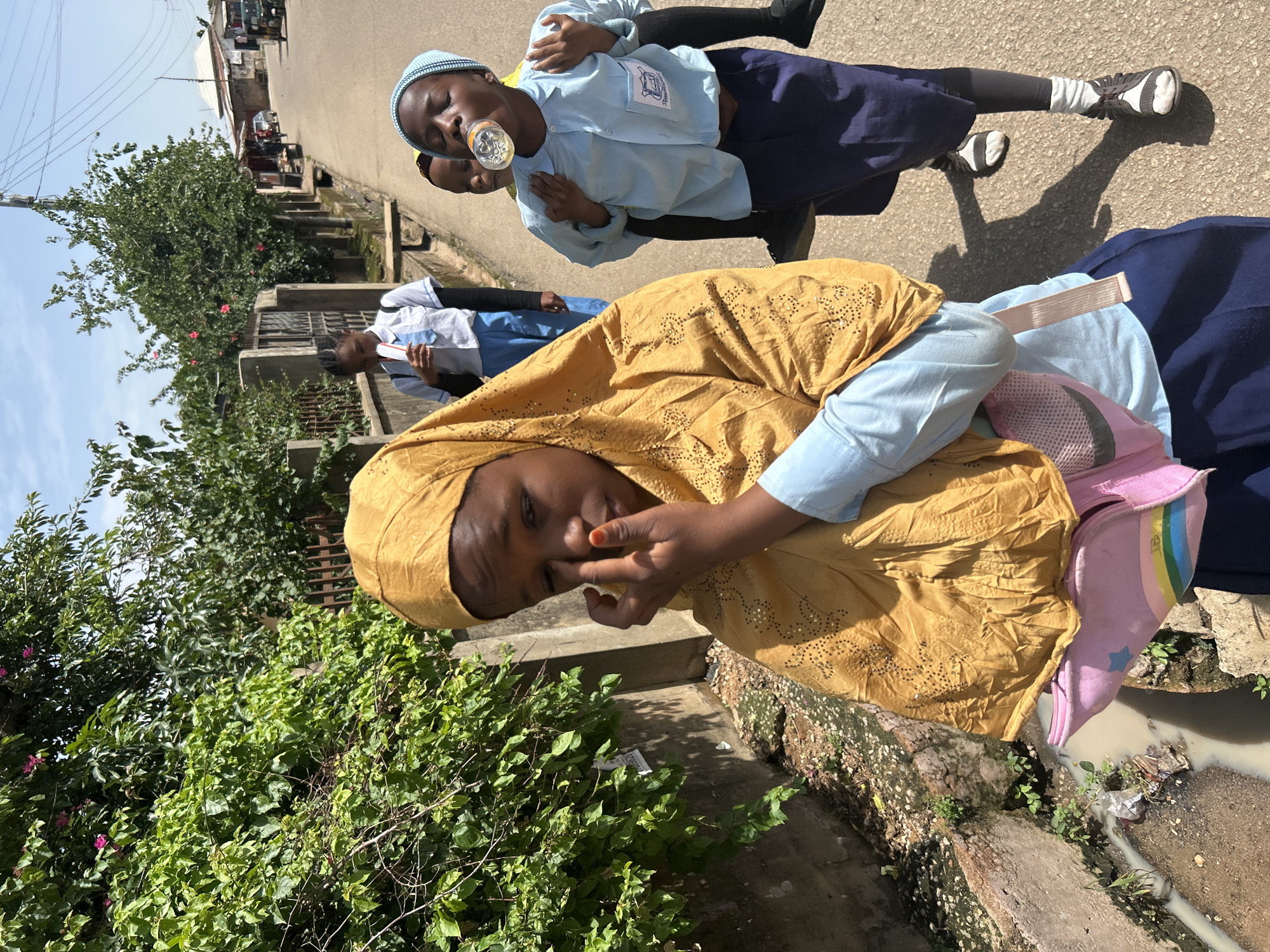 In Jos, Nigeria, four young girls walk along a paved road bordered by a concrete drainage channel. The girl in the foreground, wearing a mustard-colored hijab and a light blue shirt, touches her face. Behind her, another girl in a light blue shirt and dark blue skirt drinks from a clear bottle. A third girl, also in a light blue shirt and dark blue skirt, walks behind the bottle-drinker. The fourth girl, in a white shirt and blue pants, walks further down the road. Lush green vegetation with pink flowers grows along the side of the road, with a fence and buildings visible in the background.