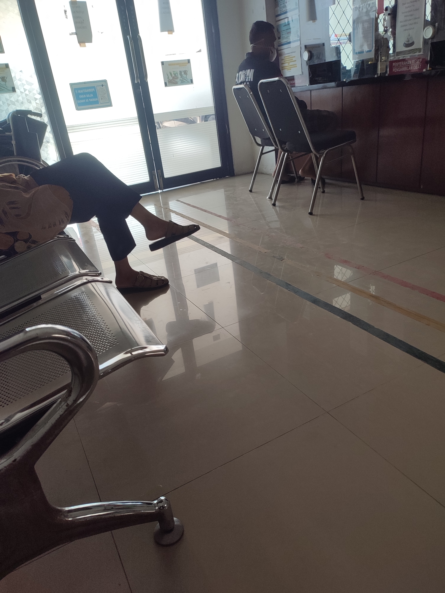 The image depicts a waiting area, likely at a medical facility, with tiled floors and fluorescent lighting. A person is seated in the foreground on a metallic bench, with their legs crossed and wearing dark pants and sandals. Their posture suggests they are waiting. In the background, another person is seated at a counter, facing away from the camera, possibly interacting with staff. The environment appears clean and functional. There are signs on the wall and counter, suggesting official information or instructions. The overall impression is one of a public waiting space.