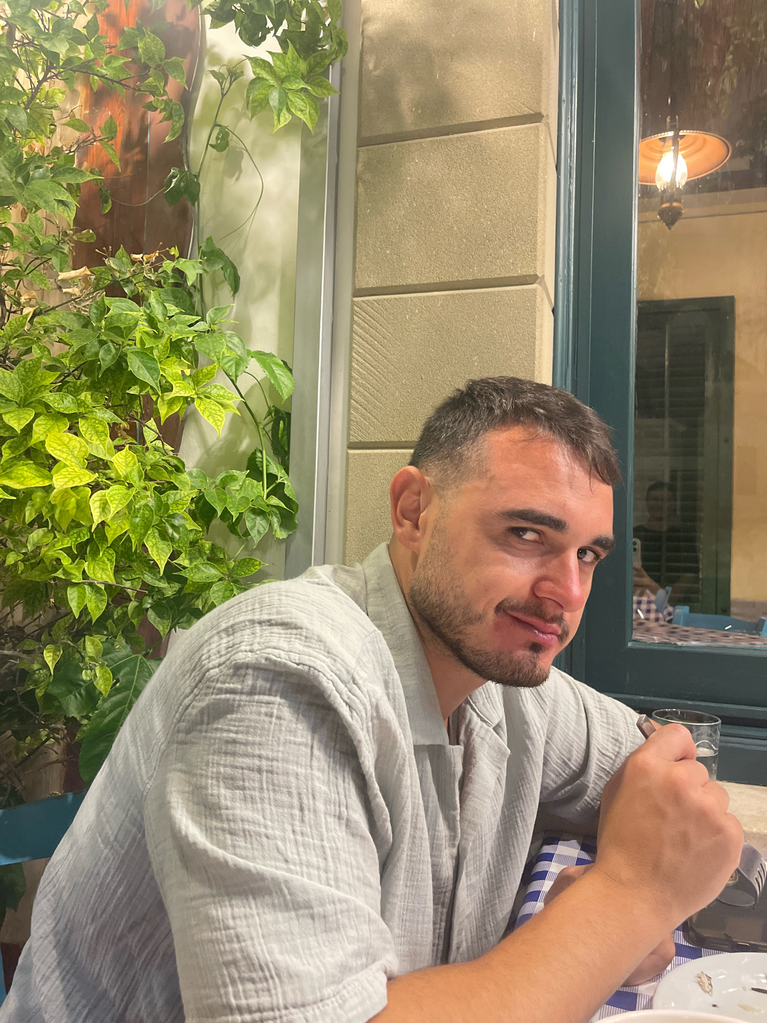 A man with short, dark hair and a beard is seated at an outdoor table, looking towards the camera. He is wearing a light gray, textured shirt. To his left, a leafy green plant is visible, with branches extending upwards and to the right. Behind him, through a window, a warm-toned hanging lamp is illuminated, and a shuttered window is visible. The man's right hand is holding a glass. The table is covered with a blue and white checkered tablecloth. The background suggests a Mediterranean setting, possibly a restaurant in Larnaca, Cyprus, given the architectural style of the building and the warm evening light.