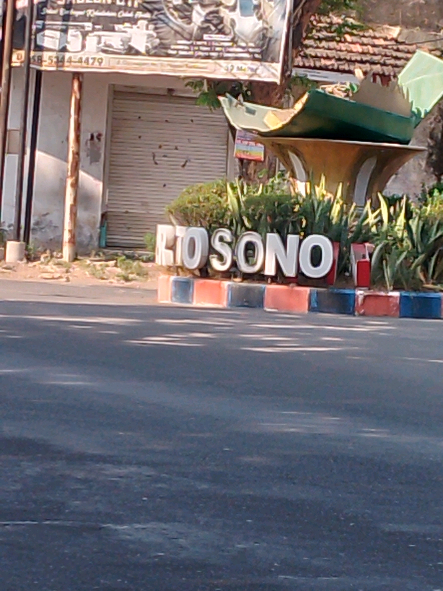 The image captures a view of a street in Kertosono, Indonesia, featuring large, three-dimensional white letters spelling out "RTO SONO". These letters are placed on a raised platform with colorful rectangular blocks along the front. Behind the letters, a lush green plant garden is visible. To the left of the letters, a building with a closed corrugated metal shutter is partially visible, along with a sign that appears to be an advertisement, displaying text and images, including a phone number. The overall setting suggests an outdoor urban environment, likely a public space or entrance to a business. The lighting indicates it is daytime, with shadows visible on the asphalt road in the foreground.