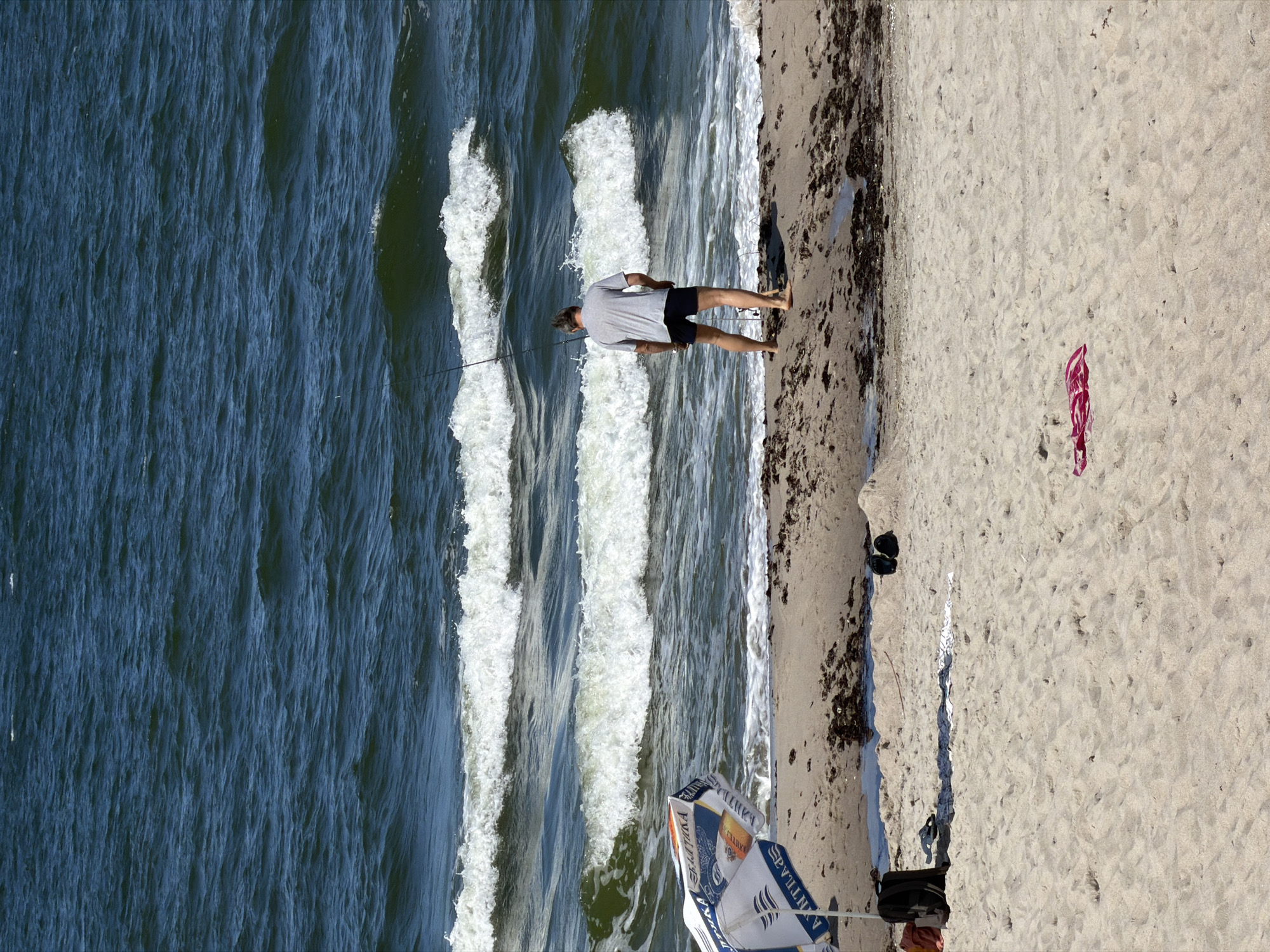 The image depicts a sunny day on a sandy beach, likely in Baltiysk, Russia, given the presence of a beach umbrella with Cyrillic lettering that appears to be a logo for "Baltika," a Russian brewery. In the middle ground, a man, viewed from behind, is actively fishing in the ocean with a fishing rod. He is wearing a grey t-shirt and dark shorts. The ocean is a deep blue with white-capped waves rolling towards the shore. In the foreground, a partially open beach umbrella is angled towards the water. Scattered on the sand are a black backpack, a pair of black flip-flops, and a red towel. The beach is composed of light-colored sand with patches of seaweed and small pools of water along the waterline. The overall scene is characteristic of a recreational day at the seaside.
