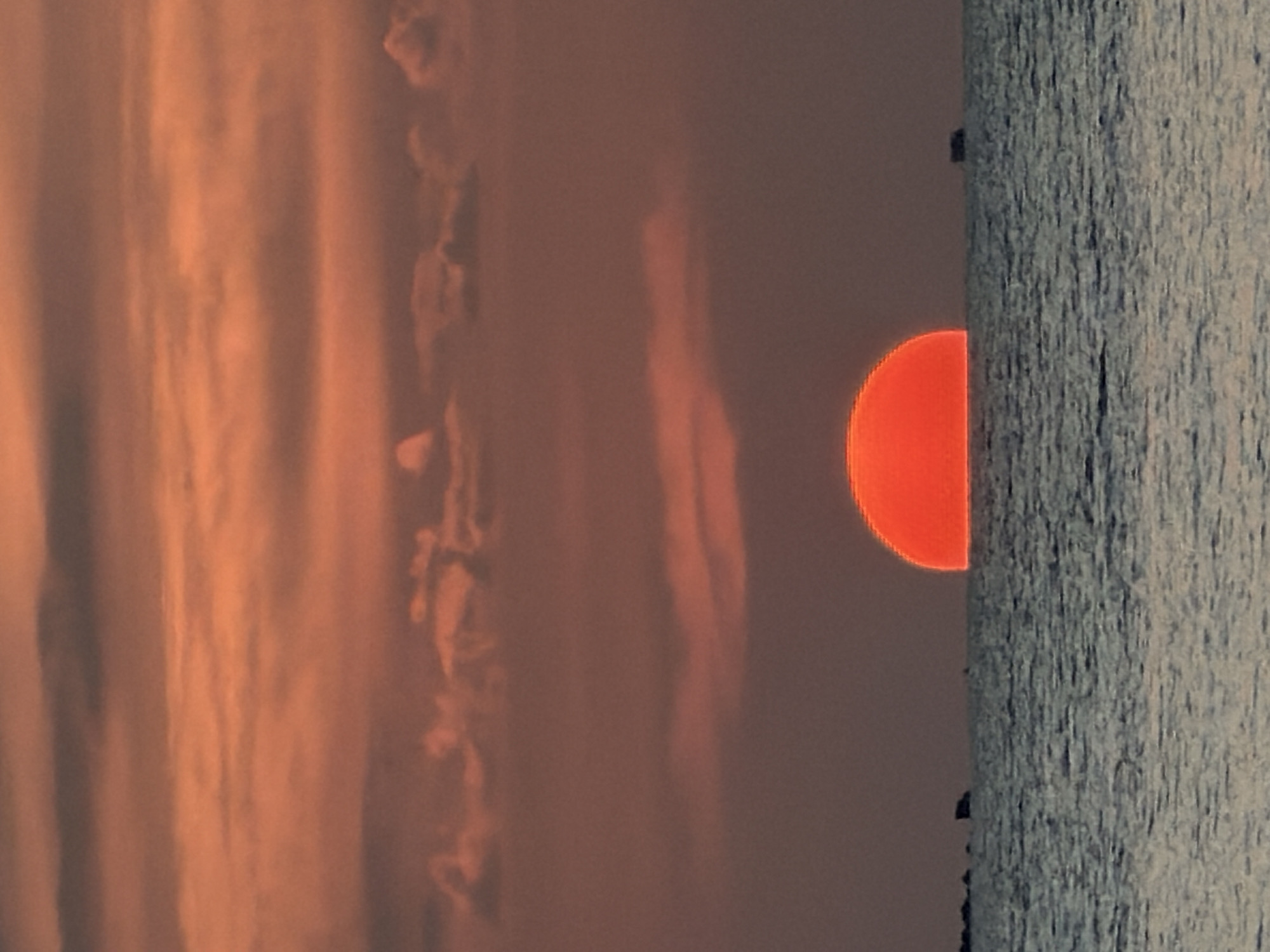 A vivid orange sun, partially obscured by the horizon, sets over a calm sea. The sky is filled with layered clouds in shades of orange and gray, creating a dramatic backdrop. The water is a deep blue, with gentle ripples reflecting the sunset's colors. In the distance, silhouetted against the horizon, are small, dark shapes that appear to be rocks or perhaps distant land formations. One of these shapes on the right side of the image resembles a small boat or structure on the water. The scene captures the tranquil beauty of a sunset over the ocean, a common sight in coastal areas like Ko Samui District, Thailand, known for its stunning beach sunsets.