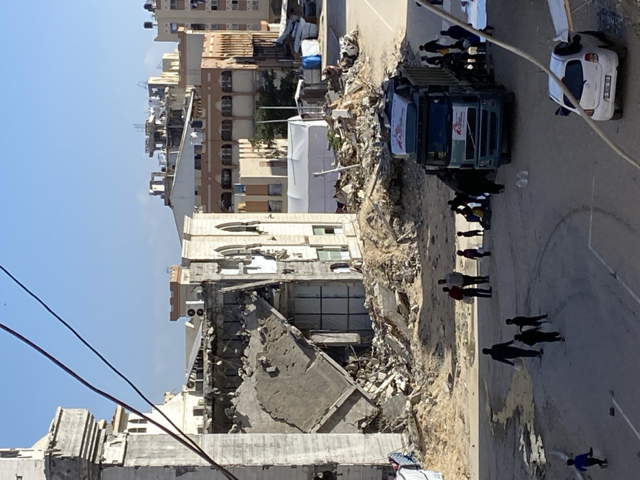 The scene depicts an outdoor urban environment in Khan Yunis, Palestinian Territory, characterized by extensive structural damage and debris under a clear blue sky.
In the left half of the image, a multi-story building is severely damaged, with large sections of its upper and middle floors collapsed, leaving a significant pile of concrete, rebar, and other building materials at its base. The remaining facade features light-colored masonry and pointed arched windows. An adjacent building, architecturally similar, also shows damage but remains partially intact.
The ground consists of asphalt road and large mounds of dirt, sand, and construction rubble. Behind these mounds, in the midground, a large white tent structure is visible, along with smaller, makeshift shelters constructed with blue tarpaulins.
Approximately 15-20 individuals, comprising adults and several children, are present on the asphalt road. In the lower right, a light blue Volvo dump truck, marked with "MSF" (Médecins Sans Frontières) logos on its cab doors and a white container on its roof rack, is stationary. Several adults are gathered near the truck, and some individuals are seen carrying white plastic containers or bags. Two adult males stand centrally on the road, one in a red shirt and the other in a grey hoodie, both holding white containers. Children are observed walking and standing among the adults. A white Nissan Micra car (license plate "P 07-XXXXX" partially visible) is parked behind and to the right of the truck. Two clear plastic water bottles are visible on the road surface near the car. Overhead utility lines traverse the upper left portion of the image.
