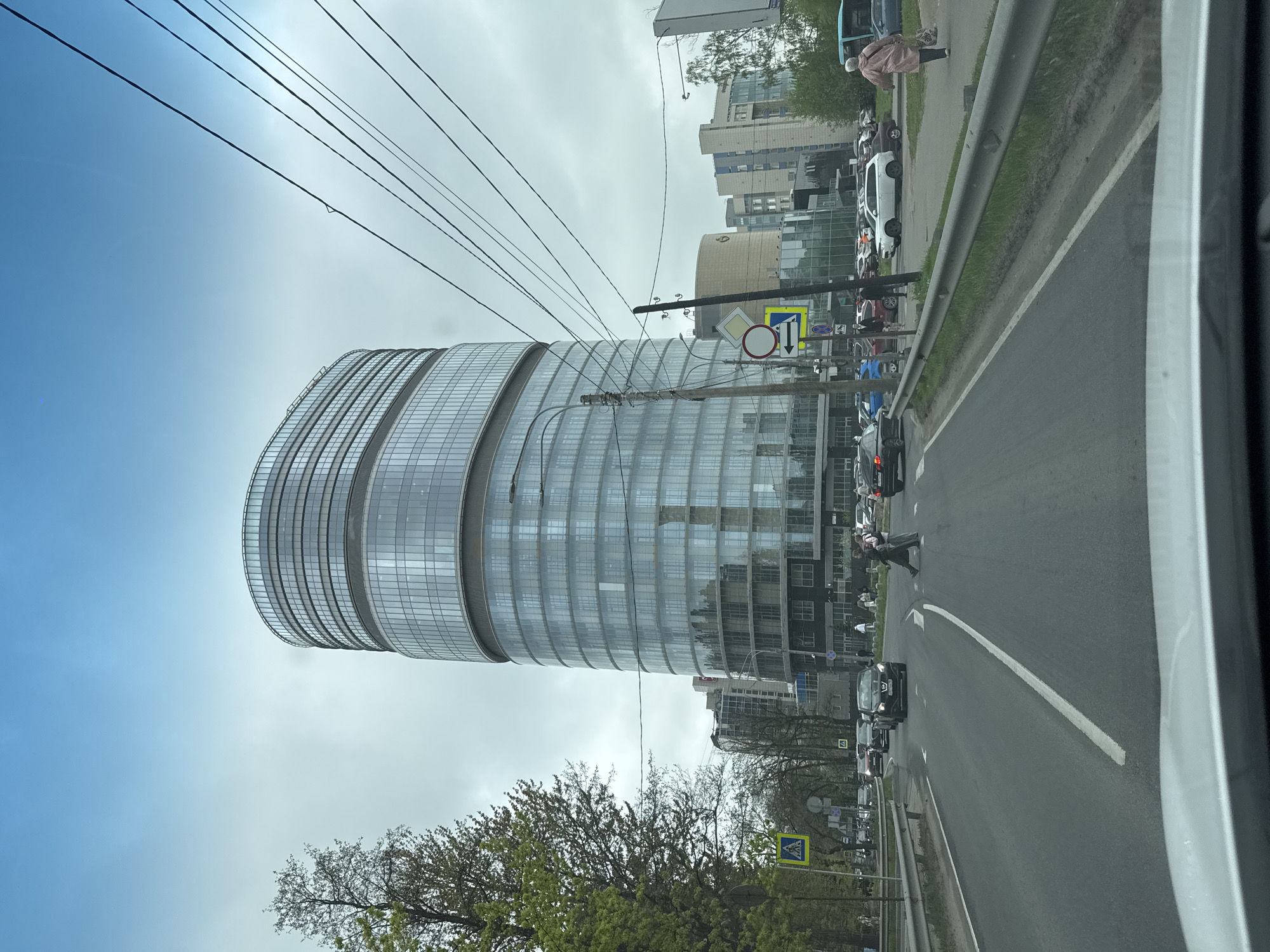 The image shows a street scene in Saint Petersburg, Russia. A modern, cylindrical glass building with horizontal banding dominates the left side of the frame. Power lines crisscross above the building. A street runs along the right side of the image with cars in both directions and a pedestrian crossing the street. Trees and other buildings are visible in the background. A street sign indicating a pedestrian crossing is visible. The sky is overcast. The photo was likely taken from inside a car.
