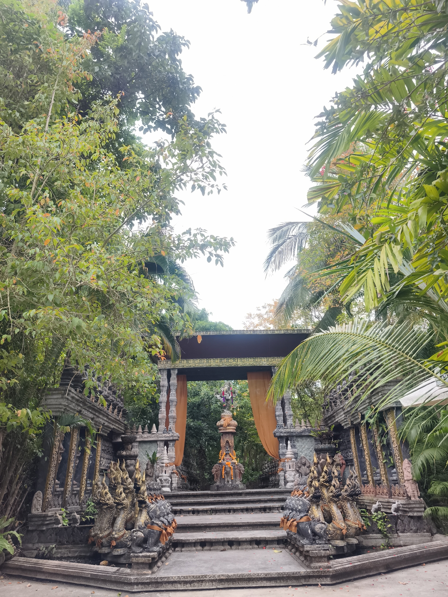 The image depicts the entrance to a Buddhist temple in Baan Lipa Noi, Thailand. The foreground features a set of concrete steps leading up to an ornate archway. At the base of the steps are sculptures of nagas (serpent deities), with details of gold accents on the sculptures. The archway is decorated with orange curtains, and a central sculpture stands beyond the archway, adorned with what appears to be a flower garland. The surrounding area is lush with tropical vegetation, including palm trees and various other plants. The sky is overcast, giving the scene a diffused light.