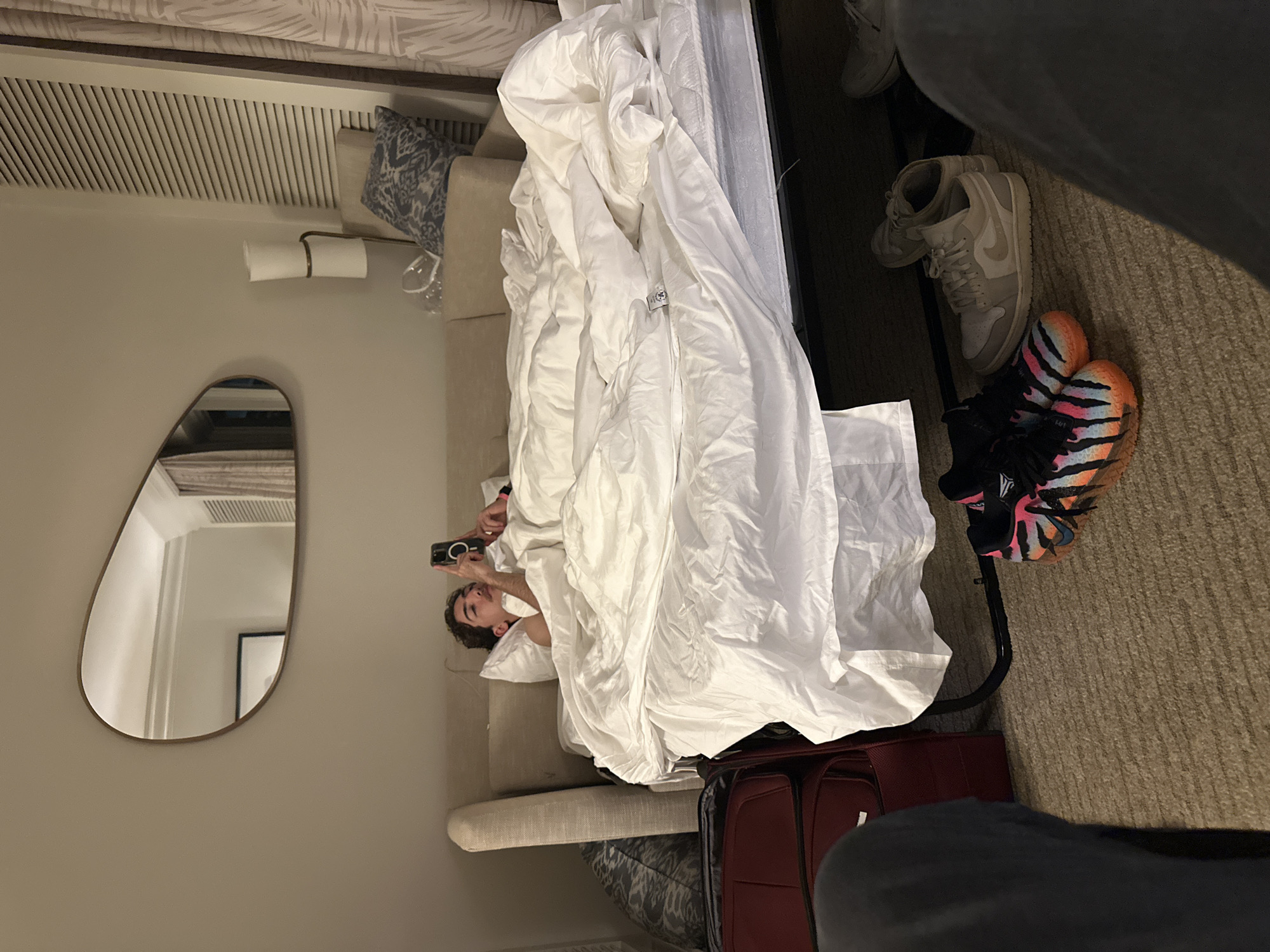 Aventura, Florida, USA. A person lies in a bed covered in white sheets, taking a photo with a smartphone. A mirror hangs on the wall. Suitcases are on the floor next to the bed. A pair of tan sneakers and a pair of orange/black sneakers are on the floor.
