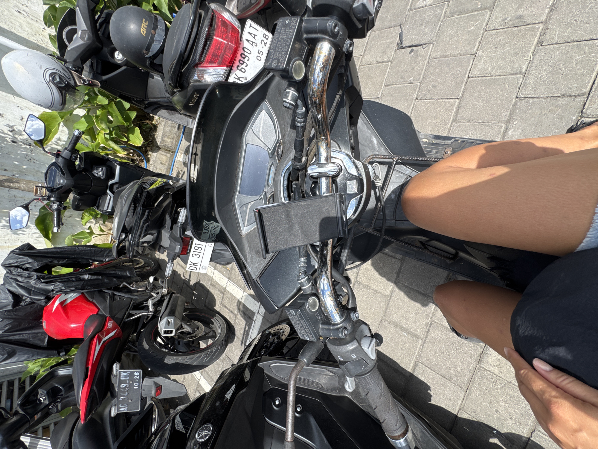 The media file shows several motorcycles parked on a paved surface. In the foreground, a person's legs are visible, indicating they are sitting on one of the motorcycles. The motorcycles are mostly black with some red and white accents. One motorcycle has a red helmet on it. The license plates are visible on some of the motorcycles, with text like "DK 3191 UK" and "AC 3449 UK". The scene appears to be outdoors, with sunlight casting shadows, suggesting it is daytime. The presence of lush green foliage behind the motorcycles hints at a tropical or sub-tropical location. The setting is likely a street or parking area in Kuta Selatan, Indonesia, given the provided location context. No specific activity or event is clearly depicted, other than the motorcycles being parked. The overall mood is casual and sunny.