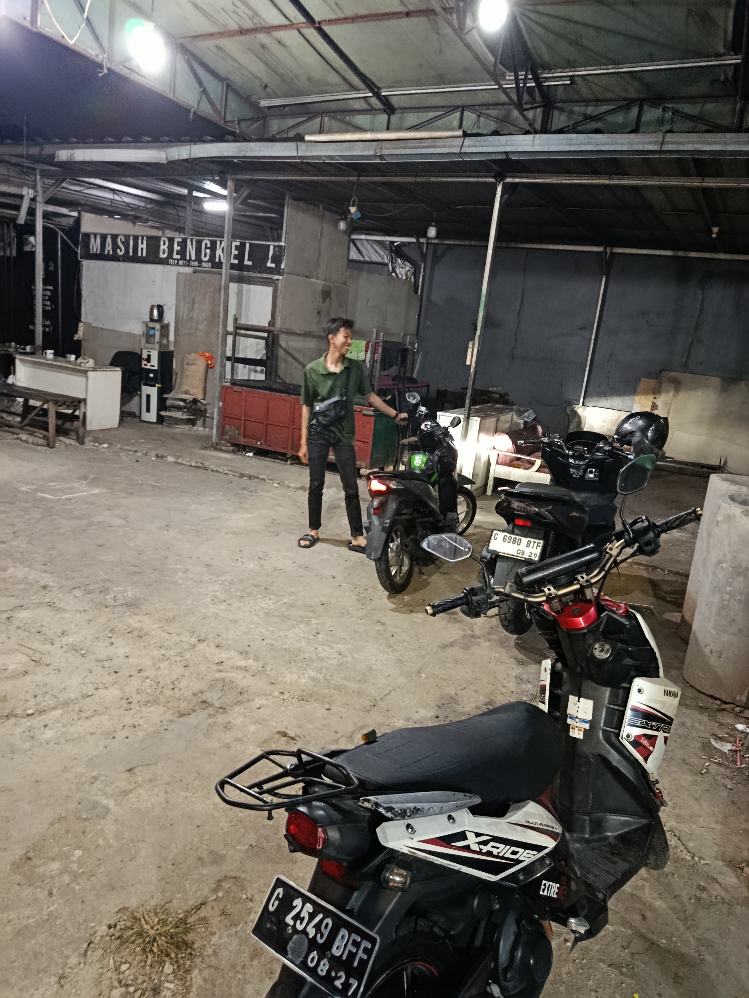 A man in a green shirt and black pants stands near several motorcycles in what appears to be a workshop or parking area. The environment suggests a commercial space, possibly a motorcycle repair shop, with concrete flooring and a covered roof supported by metal beams. A sign with Indonesian text, "MASIH BENGKEL L," is visible on a wall in the background, indicating it is still a workshop. The lighting is artificial, emanating from overhead fixtures. Several motorcycles are parked in the foreground and mid-ground, some with their lights on. The ground is dusty and uneven. In the background, there are shelves, containers, and some equipment that could be associated with a workshop. The image captures a moment in time in what is identified as Depok, Indonesia.