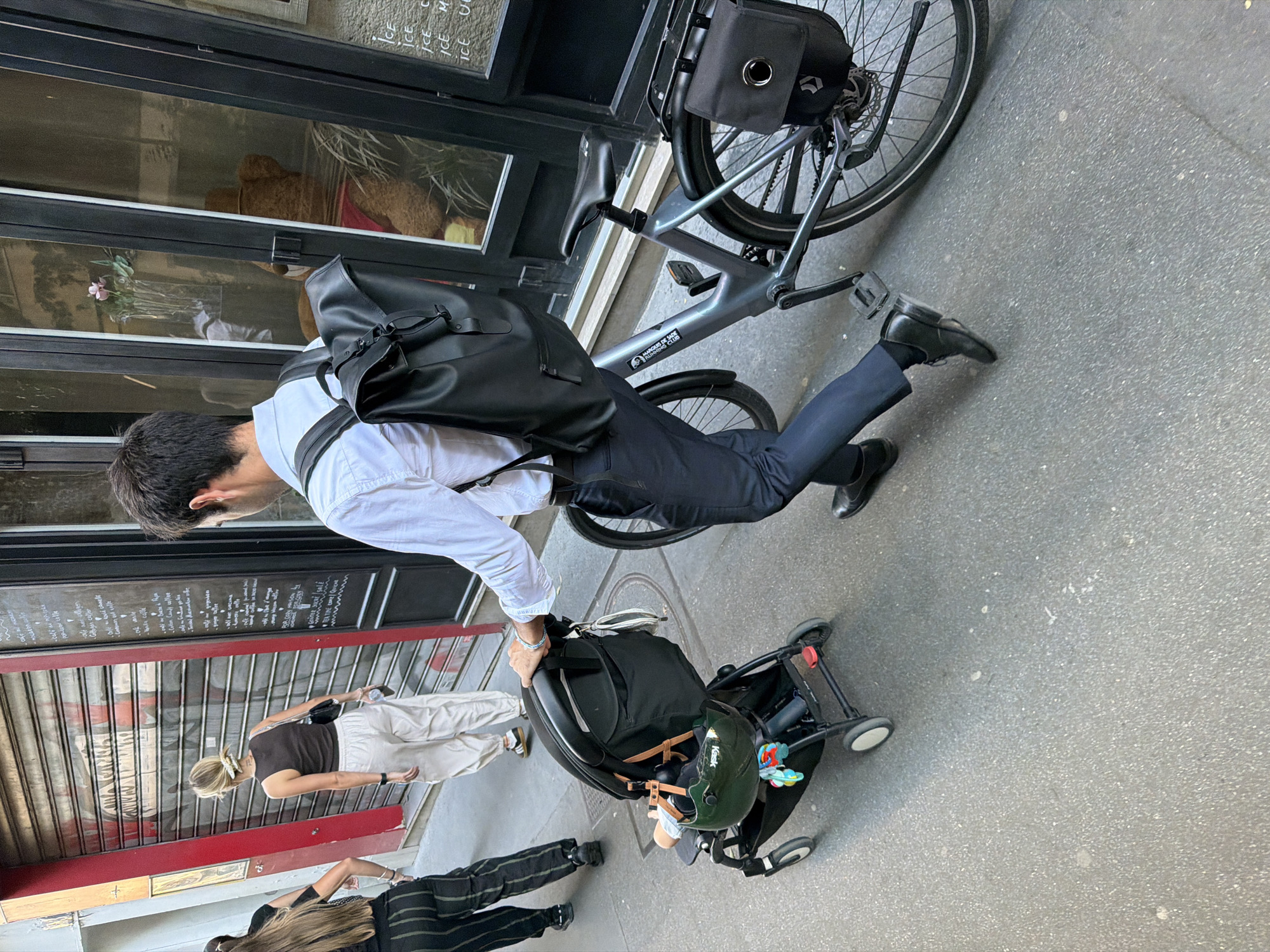 The image depicts a man in formal attire, consisting of a white shirt and dark trousers, standing on a city street. He is positioned next to a gray bicycle, with a black bag attached to its rear. The man is also holding onto a black baby stroller, where a child is wearing a green helmet.
Several other people are walking nearby, including a woman with a blonde ponytail wearing a dark top and light-colored pants, and another person wearing black-and-white striped pants.
The scene appears to be set in a commercial area, with a storefront displaying a menu or signage in French. Notably, the words "MARQUIS DE" are visible on the bicycle. Based on this inscription, the presence of French signage, and the architectural style, the location can be inferred to be in the city of Paris, France.