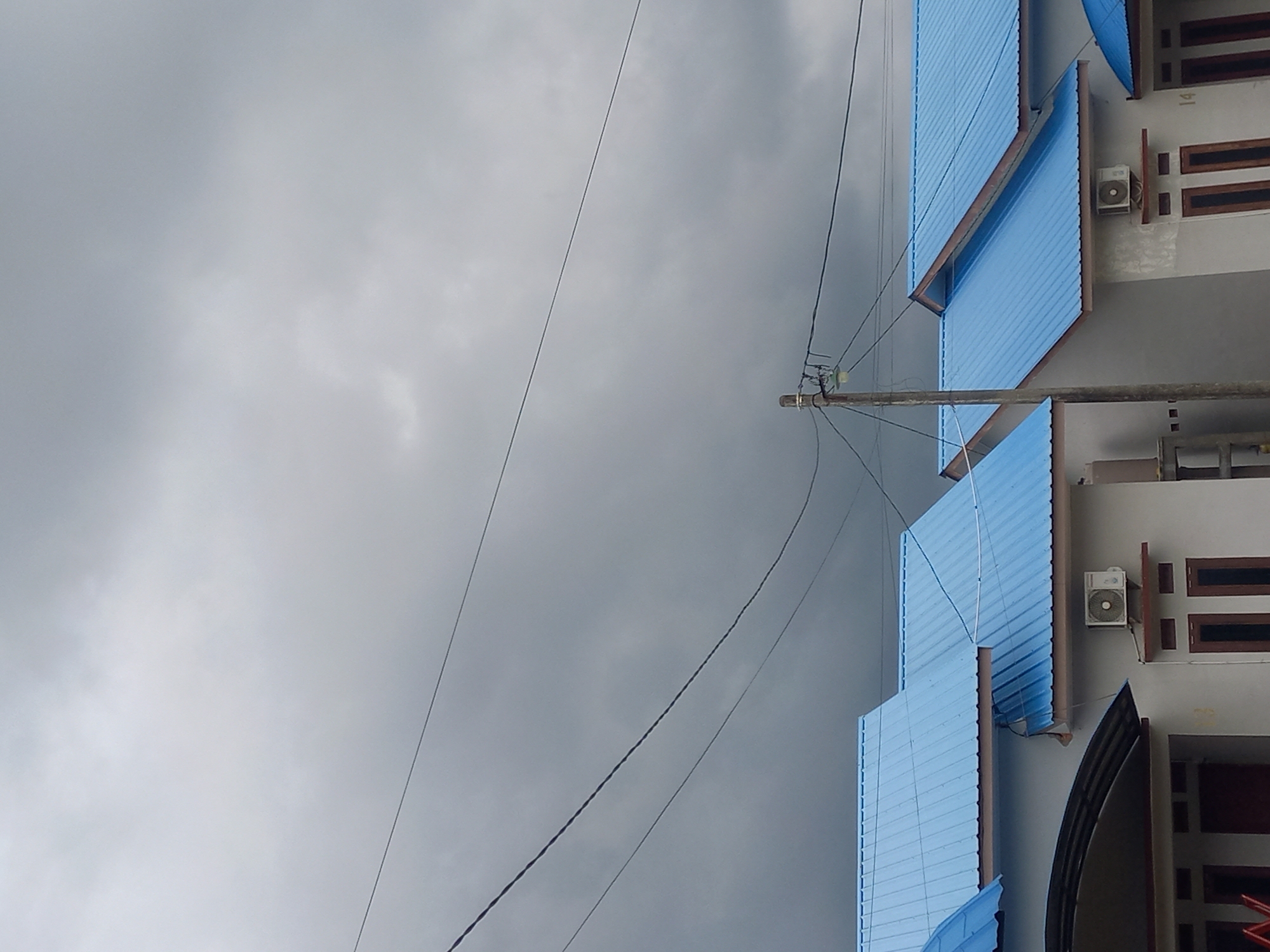 The image displays an overcast daytime scene in Puloblang, Indonesia, featuring multiple buildings, a utility pole, and numerous overhead wires against a gray sky.
On the right side of the frame, several multi-story buildings with light gray or off-white walls are visible. These buildings are topped with bright blue, corrugated metal roofs, some of which extend outwards as awnings. Dark brown, rectangular windows are set into the building walls. At least two outdoor air conditioning units are mounted on the walls; one is positioned between two windows on an upper floor, and another is visible on a lower floor. Faint text, specifically the number "14", can be seen on the wall of one building, and a partial "23" is barely discernible on a lower section.
A wooden utility pole stands vertically on the right side, carrying insulators and a cluster of power lines. Numerous black cables and wires stretch horizontally across the frame, originating from or passing by the utility pole, creating a complex network against the sky.
The left side and upper portion of the image are dominated by a dense, gray, overcast sky, indicating cloudy weather. There is no human activity depicted in the static scene.