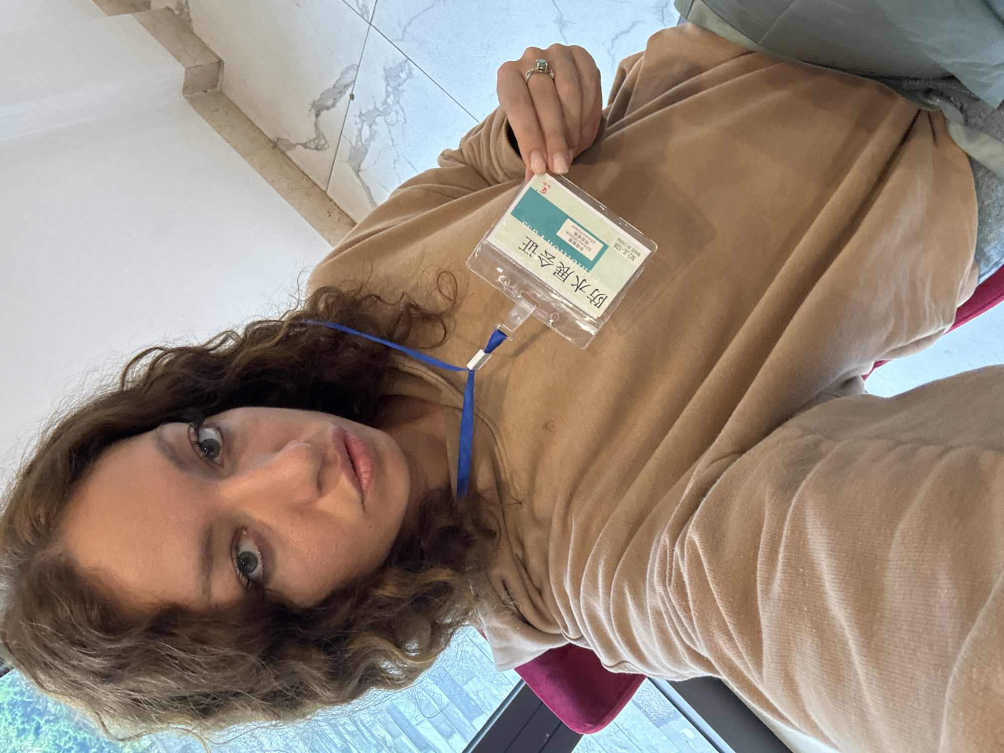 A woman with curly brown hair and fair skin is holding up a badge in a selfie. She is wearing a tan-colored hooded sweatshirt and a ring on her left hand. The badge is clear and has a blue lanyard. The text on the badge is in Chinese. In the background, a marble floor and a window with a view of trees can be seen. The location is Binh Lu, Vietnam, based on the Chinese text that translates to Waterproof Exhibition Certificate.