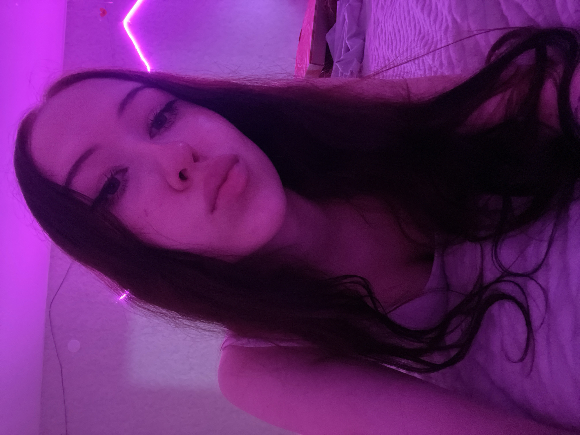 The image is a close-up selfie of a young woman lying on a bed. She has long, dark hair and is looking directly at the camera. Her face is illuminated by a bright pink neon light, which also casts a pink glow on the surrounding environment. The background appears to be a bedroom with a textured wall and a bed covered in a textured comforter. There is a pink neon light strip visible in the upper right corner, forming a geometric shape. A pink box is on the bed to the right of the woman. The overall impression is one of a casual, personal moment captured in a dimly lit, vibrantly colored room. There is no information in the image to suggest a specific location in Votkinsk, Russia.