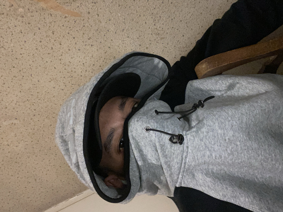 A person is looking into the camera while wearing a grey hoodie with a black lining. The hoodie is pulled tightly around the face, obscuring most facial features except for one eye and part of the nose and ear. The person is wearing black pants, and a wooden chair is visible on the right side. The background is a mottled beige surface.