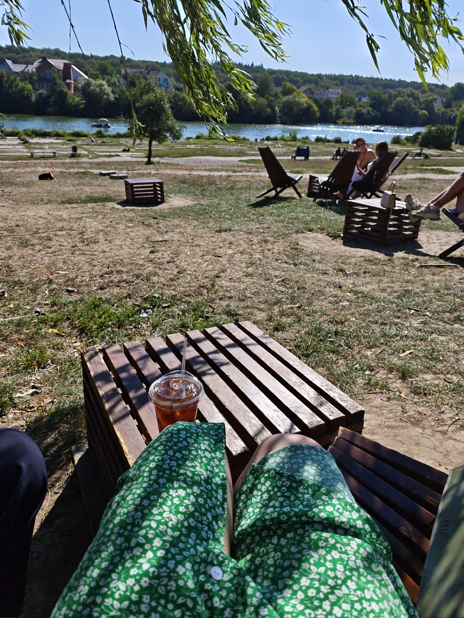 The image captures a sunlit outdoor seating area near a body of water, possibly the Southern Bug river, in Vinnytsia, Ukraine. In the foreground, a person wearing a green floral dress is seated on a wooden bench with a clear plastic cup of iced beverage and a straw on a low wooden table made of pallets. To the left, another person's legs, clad in dark trousers, are visible. In the midground, several wooden lounge chairs are occupied by individuals, some of whom appear to be relaxing and conversing. One person is wearing a white top, and another's legs are visible in sneakers. Further back, wooden pallet tables and chairs are scattered across a dry, grassy area. A small boat is visible on the water, and houses and trees line the opposite bank. The sky is clear and blue. The overall atmosphere suggests a casual outdoor recreational space.