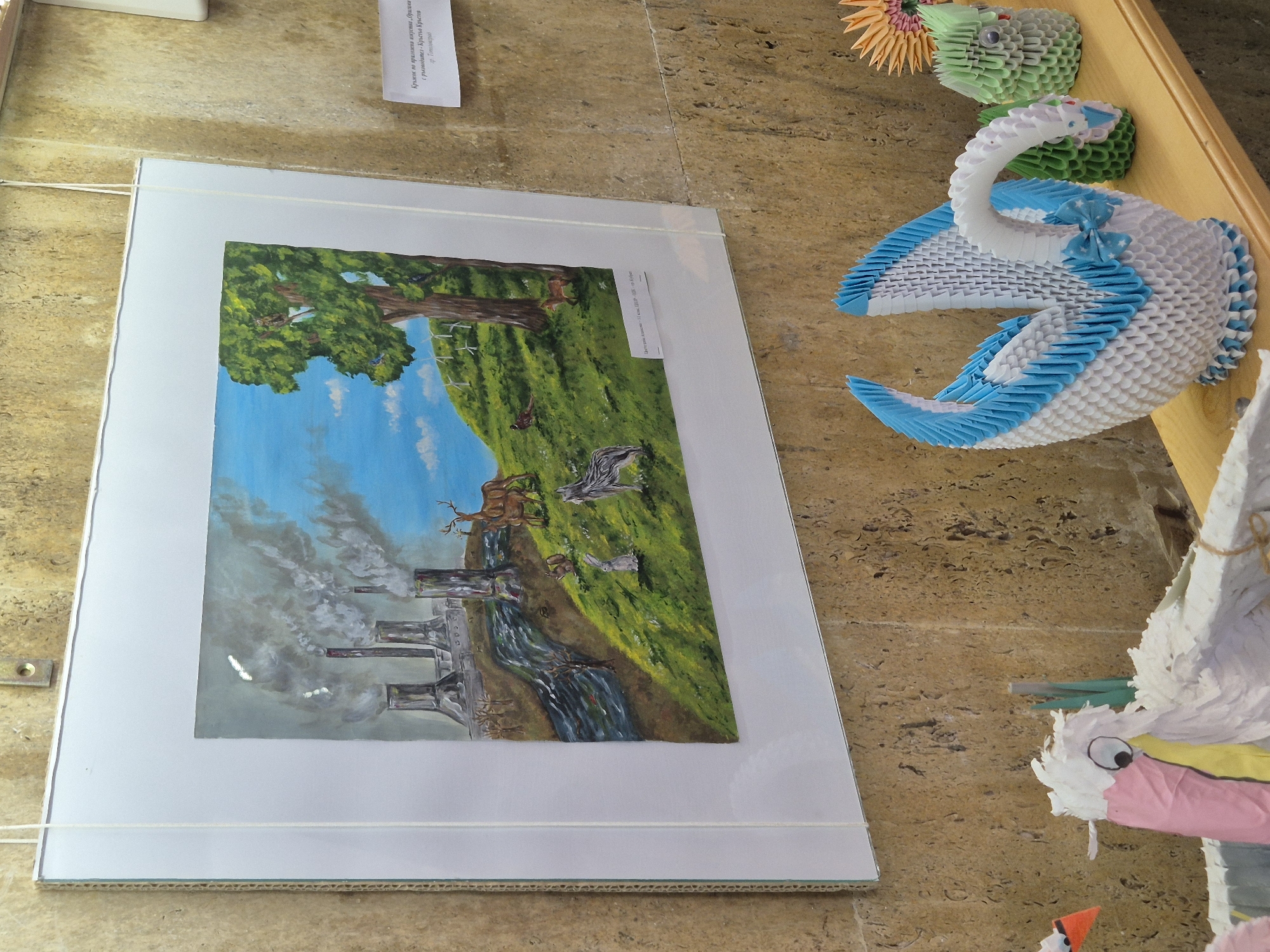 The image captures an art exhibit. A framed painting shows a transition from industrial pollution on the left to nature with trees, animals, and a blue sky on the right, suggesting environmental contrast. To the right of the painting are origami figures, including a swan. The image is set against a textured background. The scene is possibly at or related to the Lake Srebarna nature reserve.
